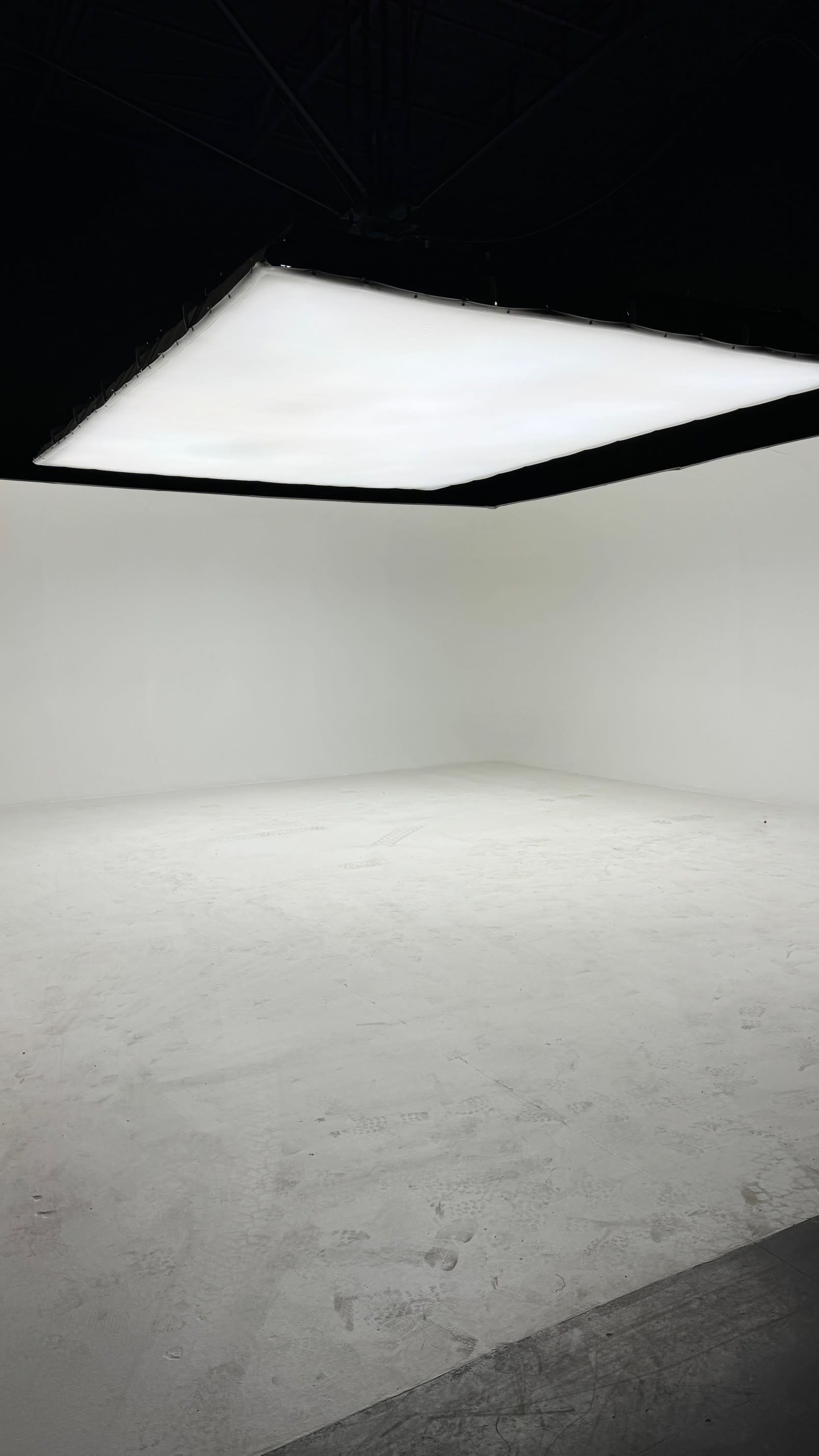 The image shows a spacious, minimalist room with white walls, a white floor, and a large rectangular light fixture hanging from the ceiling.