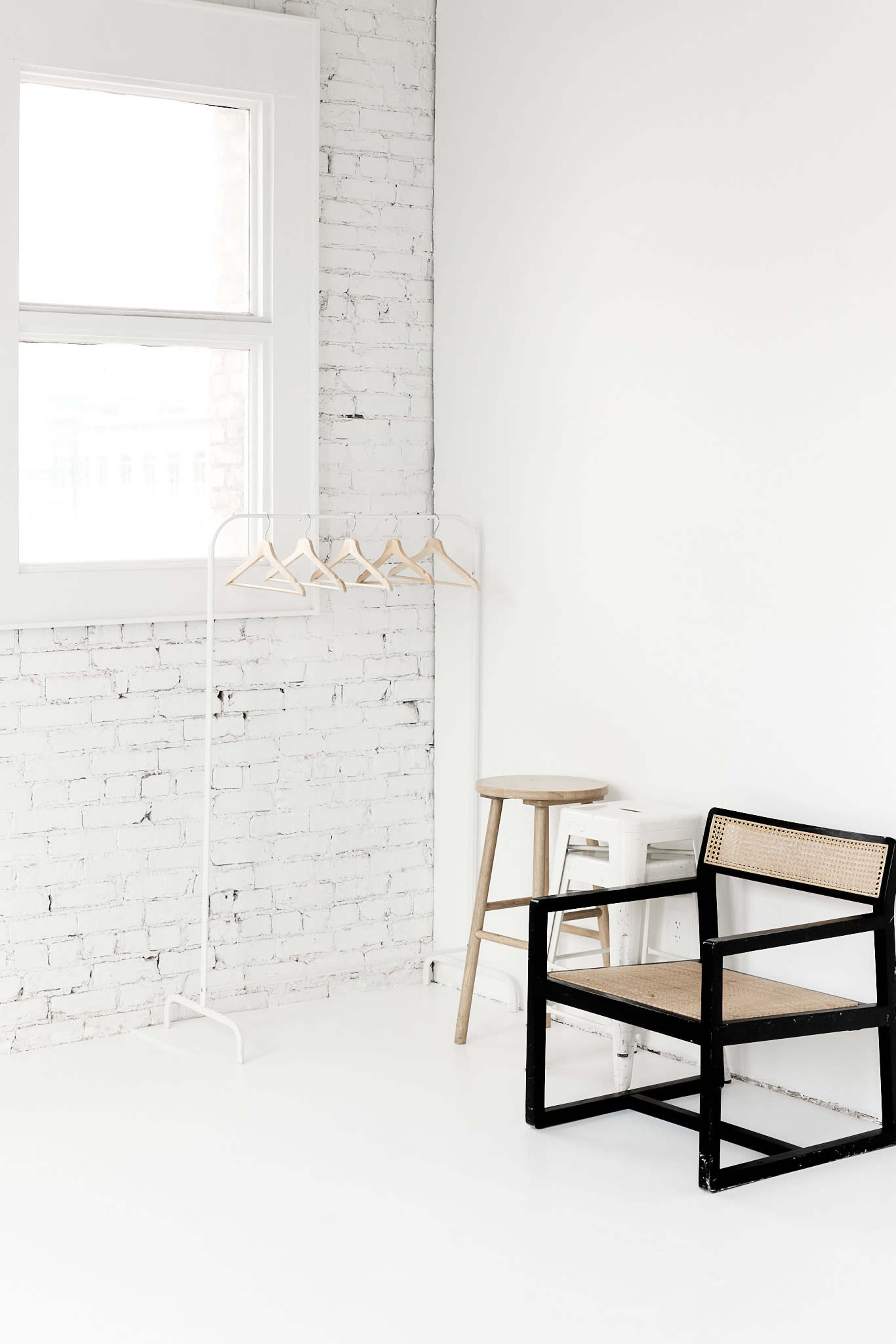 The image shows a minimalist interior with a white brick wall, a clothing rack with empty hangers, a wooden stool, and a black and cane chair.