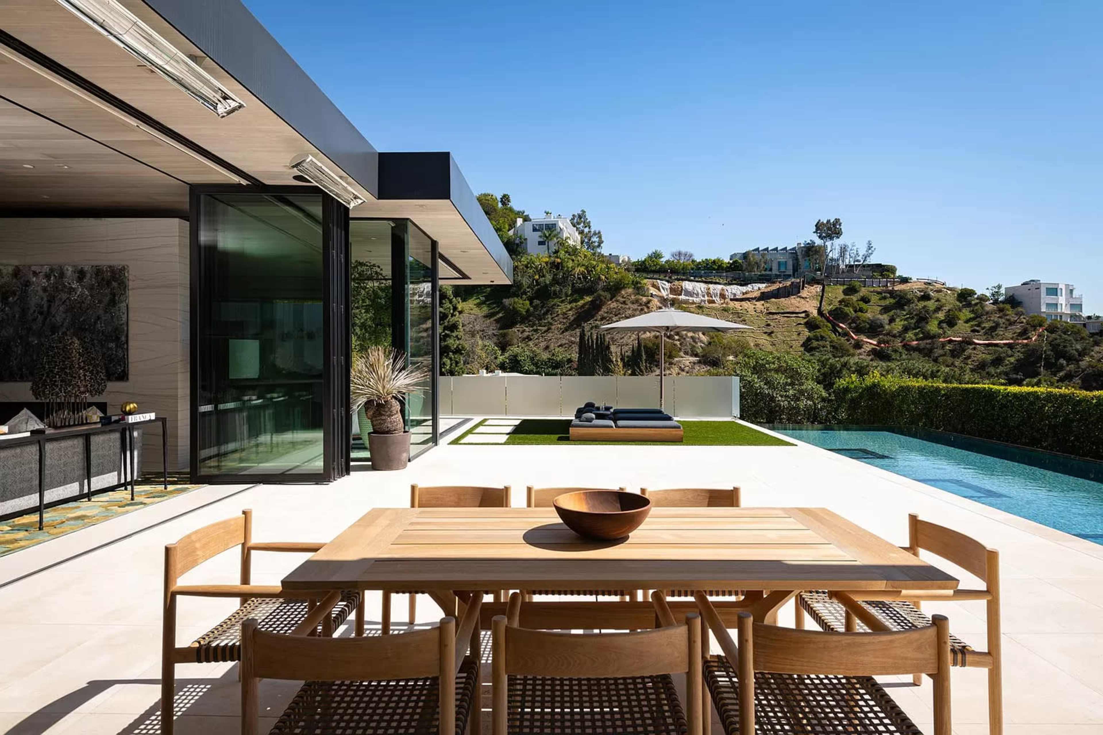 A modern outdoor patio features a wooden dining table with chairs, a landscaped hillside in the background, a pool on one side, and an umbrella shading a lounge area.