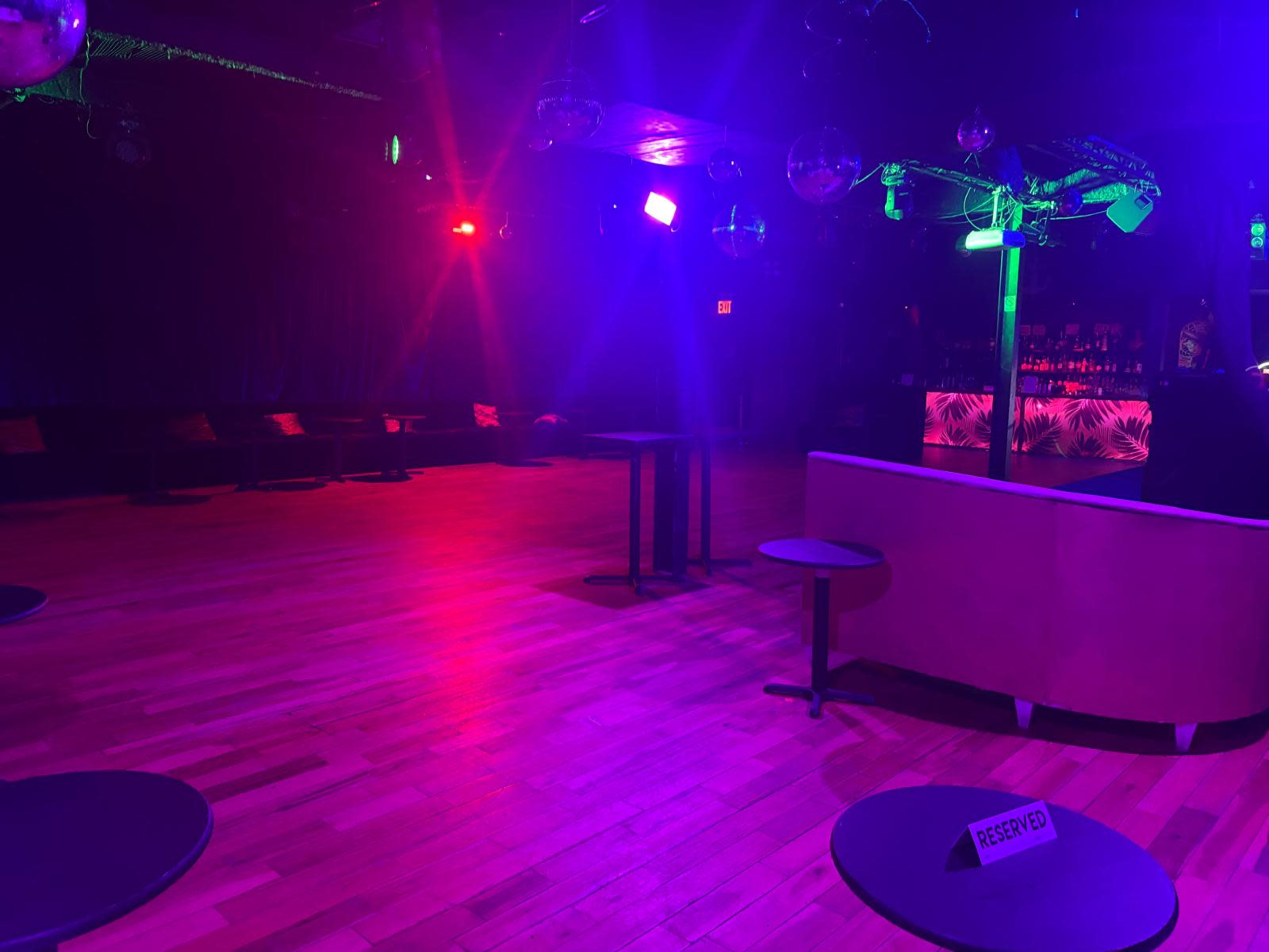 The image shows an empty nightclub with colorful lighting, featuring wooden floors and reserved tables.