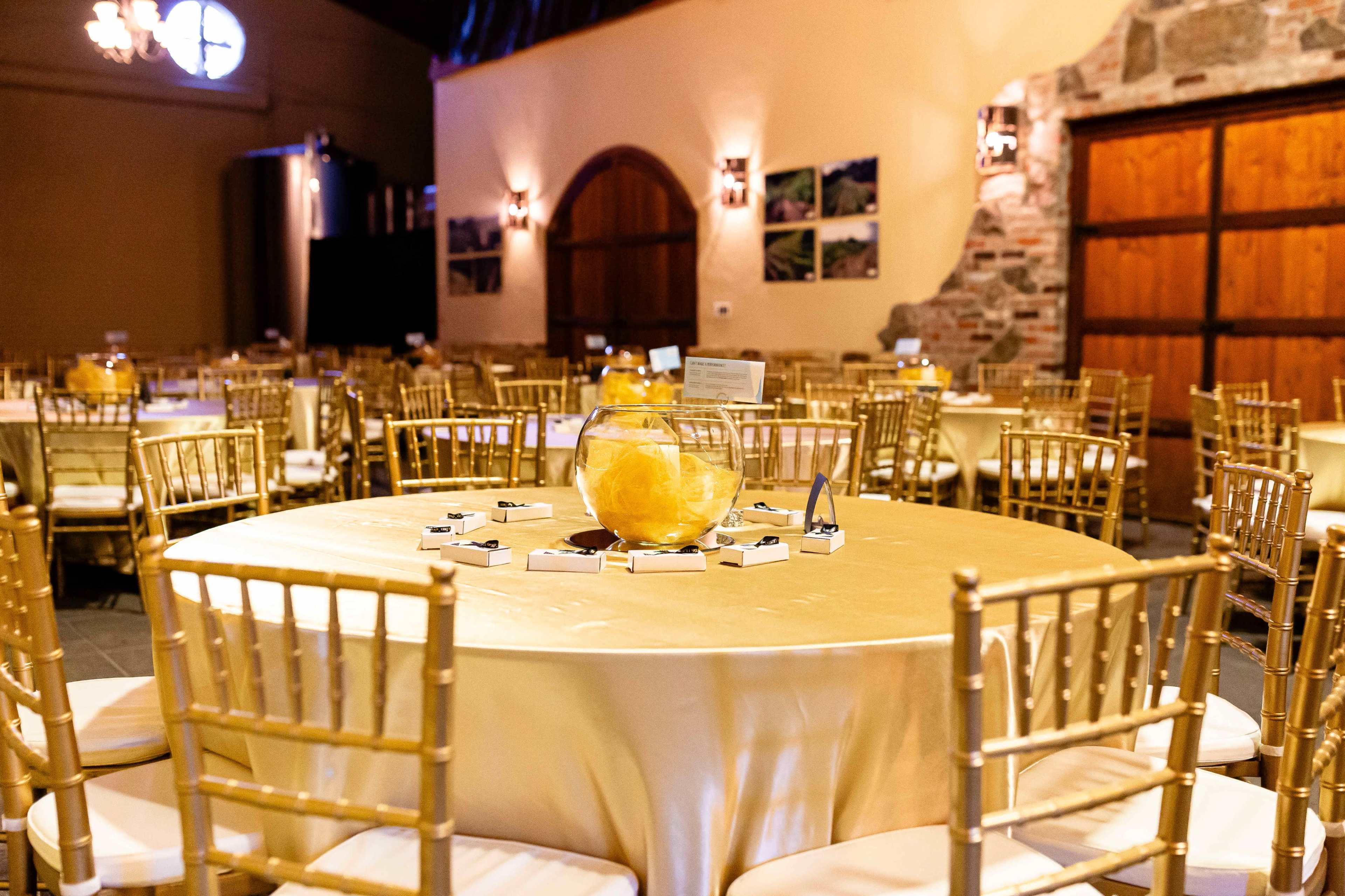 A large event hall with round tables covered in gold tablecloths, each adorned with a glass bowl of yellow decor and surrounded by gold chiavari chairs.