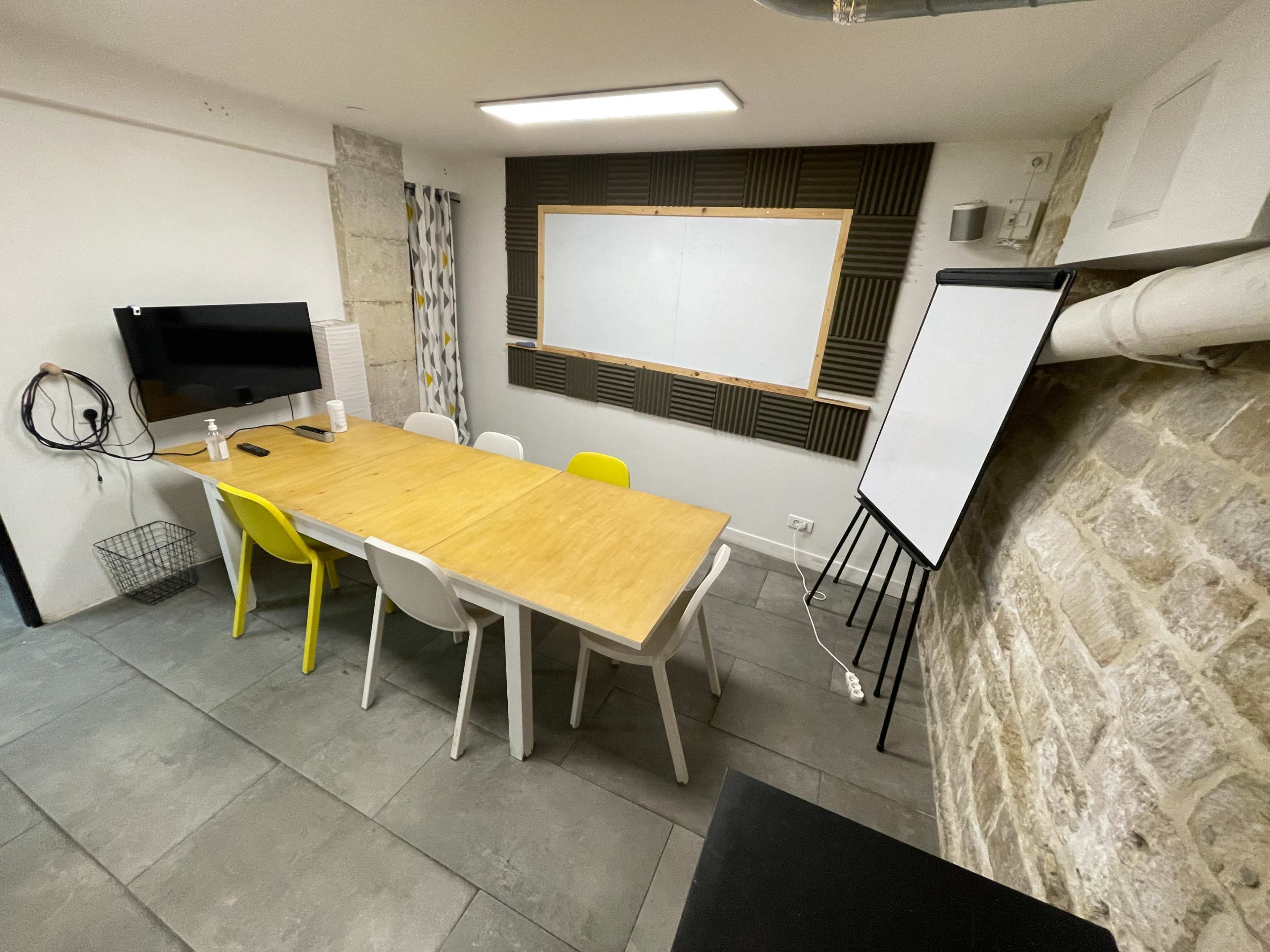 A meeting room features a long wooden table surrounded by colorful chairs, a whiteboard, a television on the wall, and exposed stone elements.