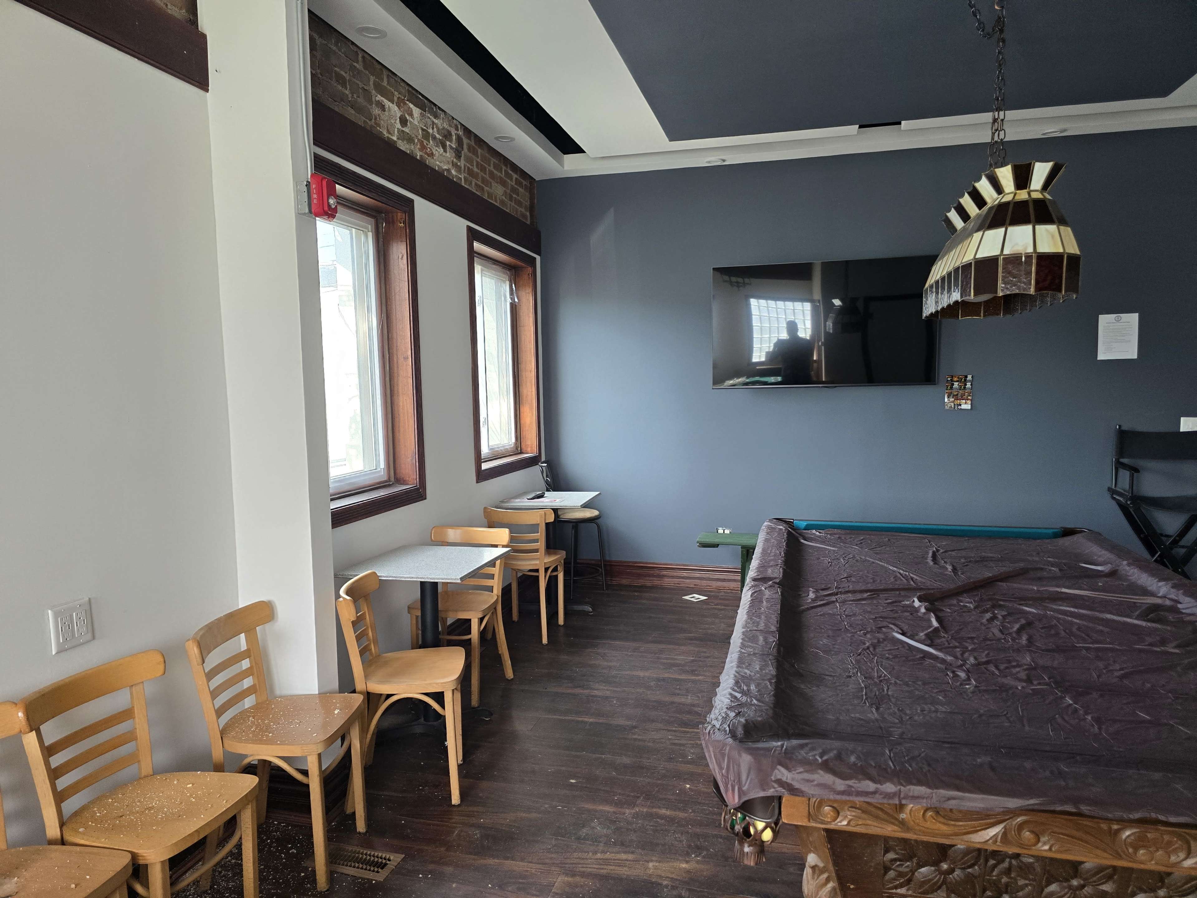 The image shows a room with a billiard table, several wooden chairs and small tables, and windows allowing natural light, alongside a wall-mounted television.