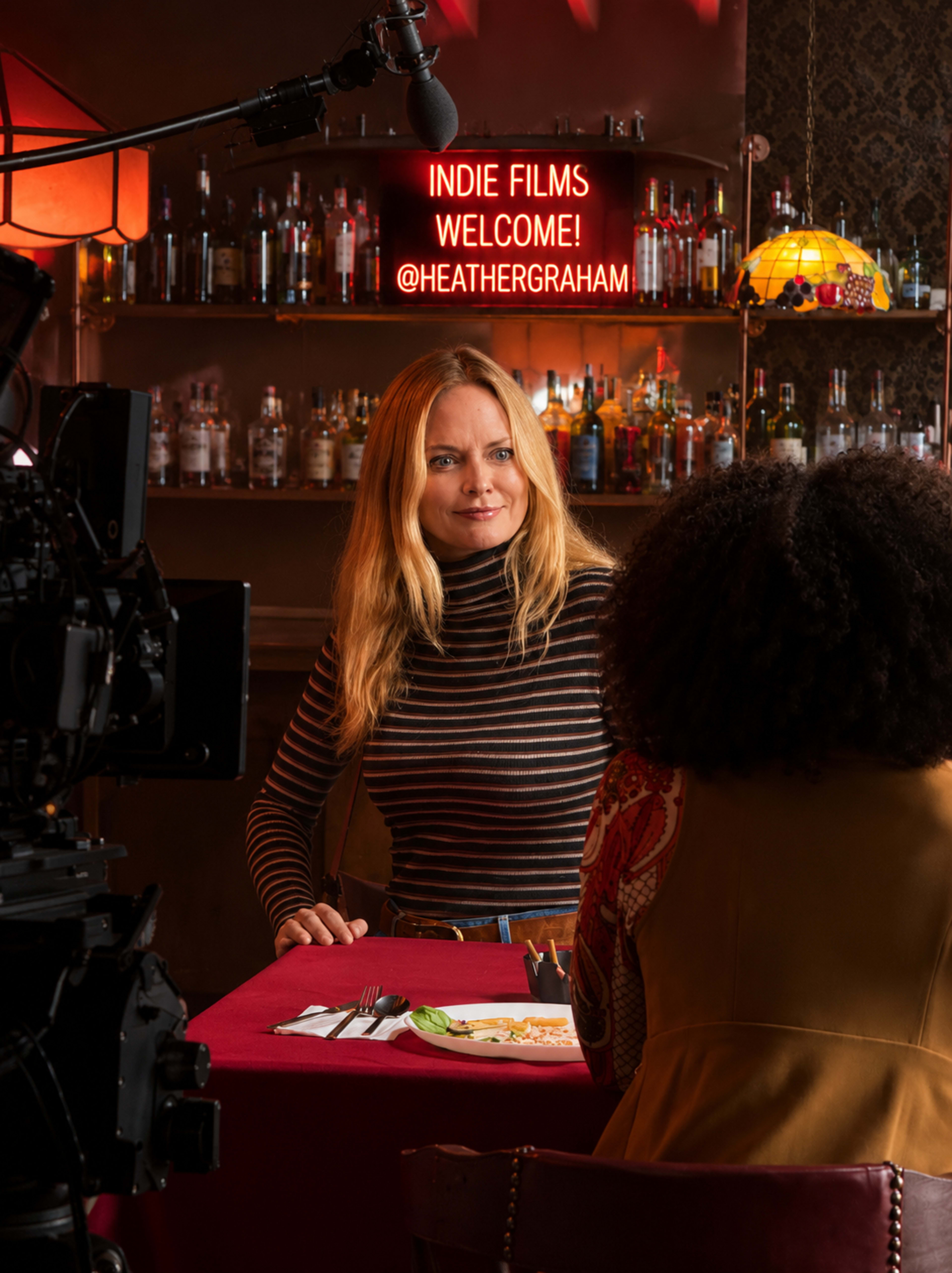 A woman sits at a table in a film set restaurant, with a camera and crew visible in the foreground and a neon sign in the background that says "INDIE FILMS WELCOME!"