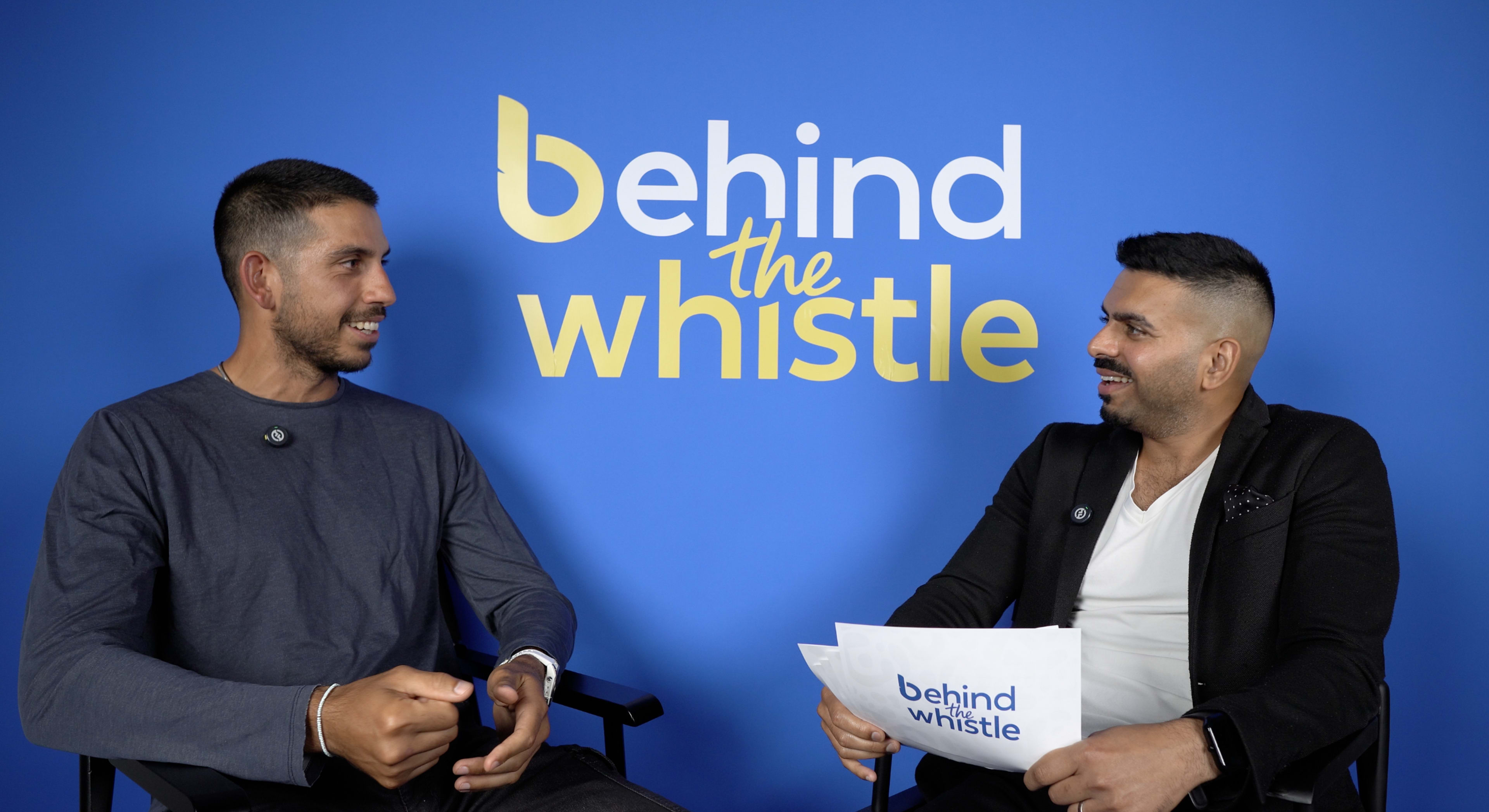 Two men sit across from each other in a studio setting, engaging in conversation against a blue backdrop featuring the text "behind the whistle."