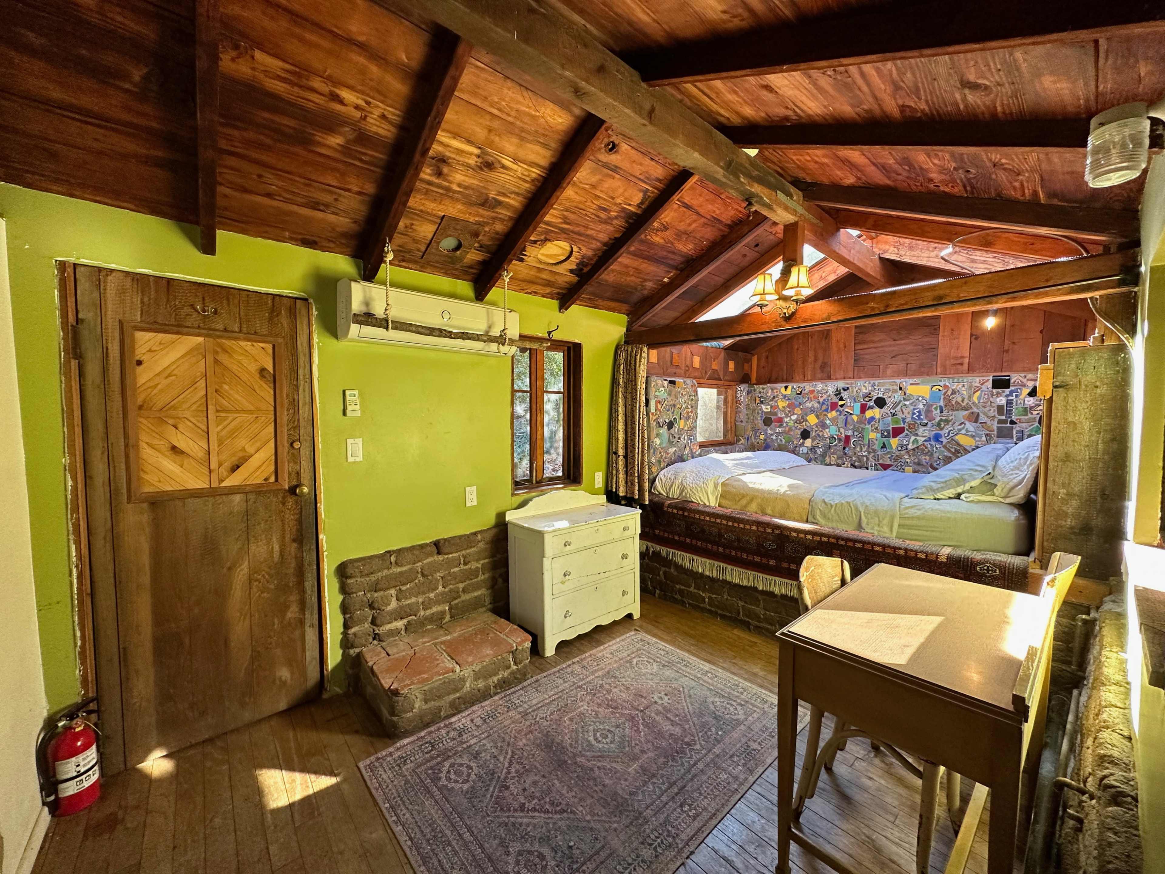 A cozy room with a wooden ceiling, a double bed with a colorful mural behind it, a small desk, and a door leading outside.