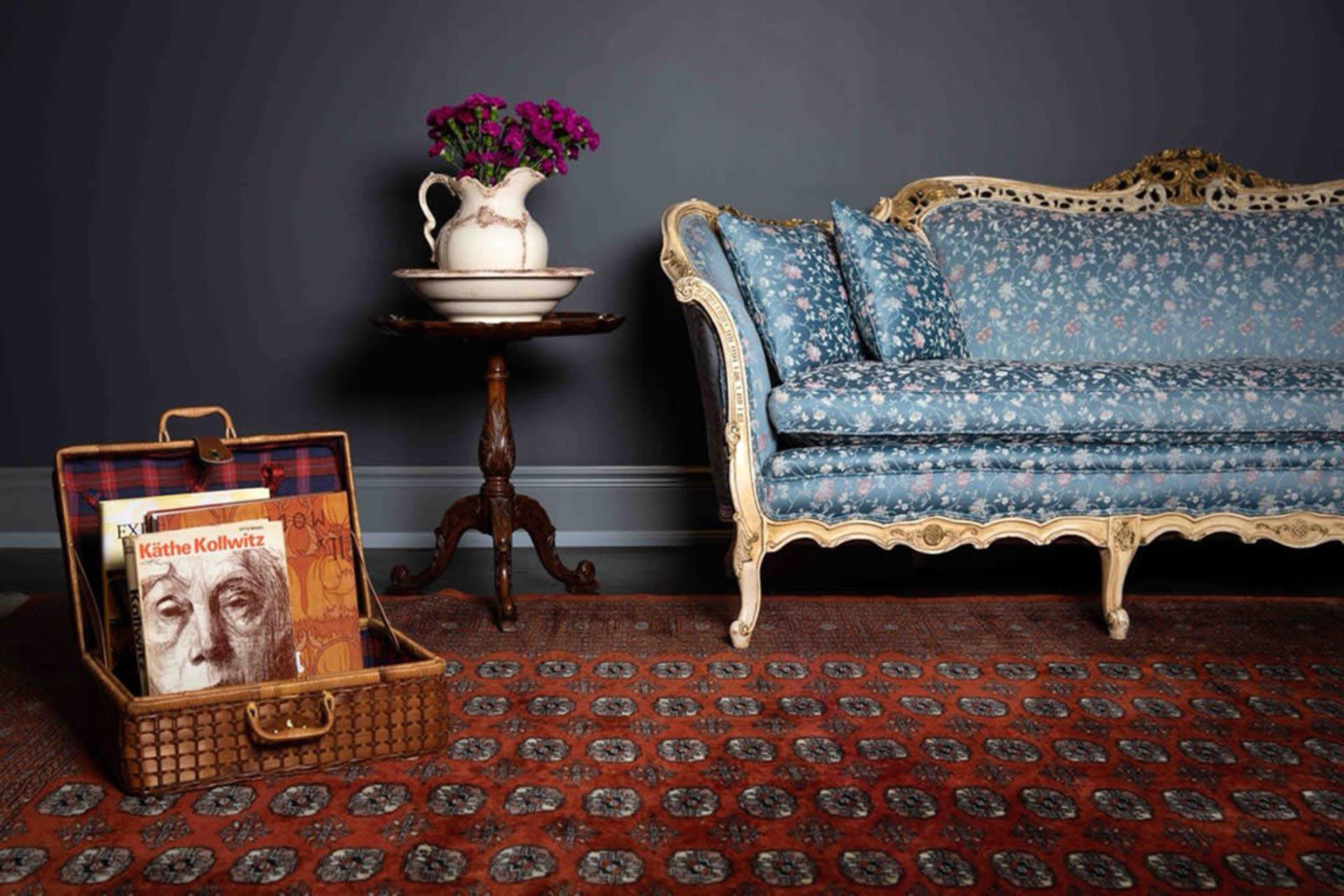 The image features a vintage blue floral couch next to a small round table with a pitcher and a pot of flowers, with a wicker basket containing art books on the floor.