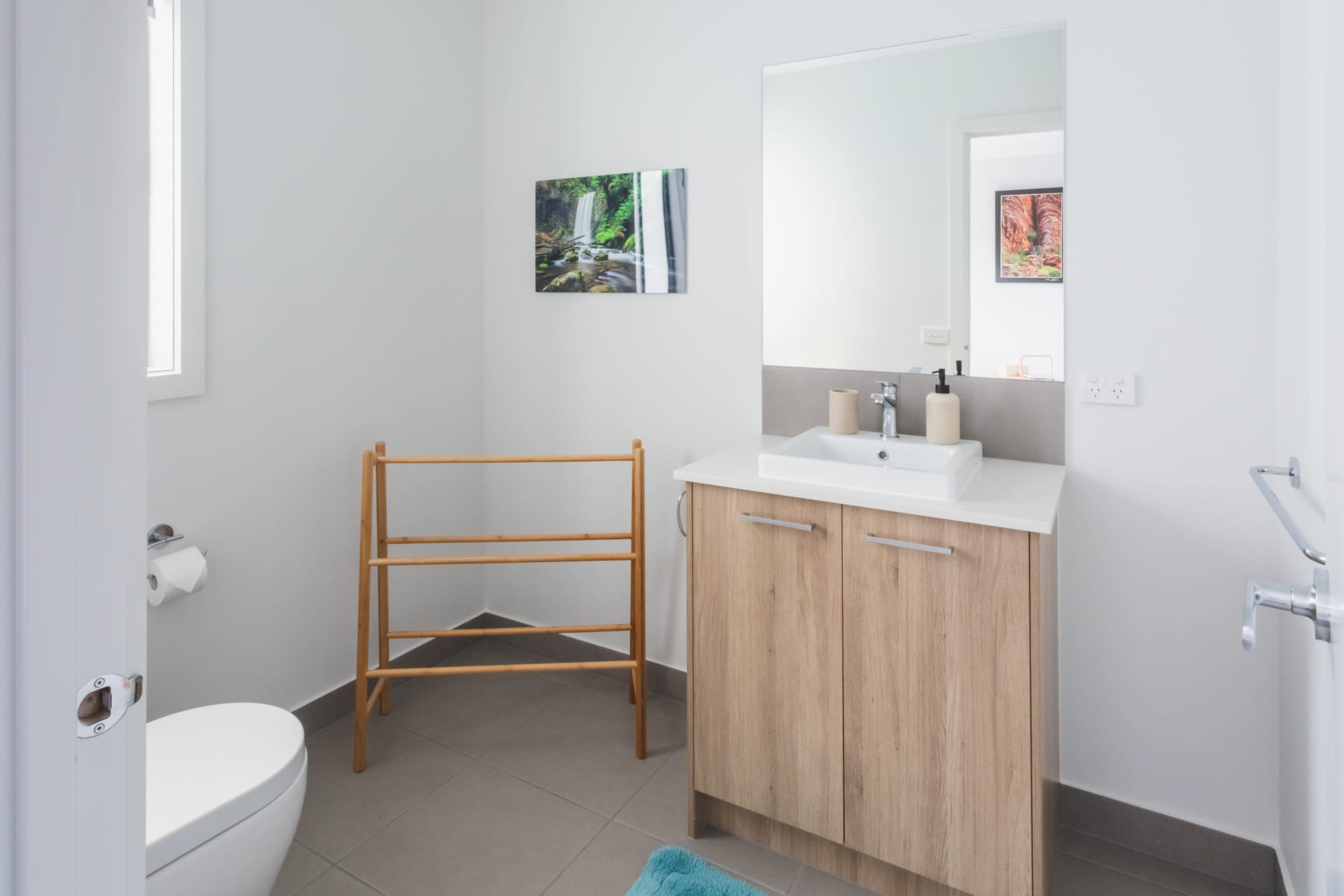 A modern bathroom features a wooden cabinet with a sink, a toilet, a towel rack, and a framed nature picture on the wall.