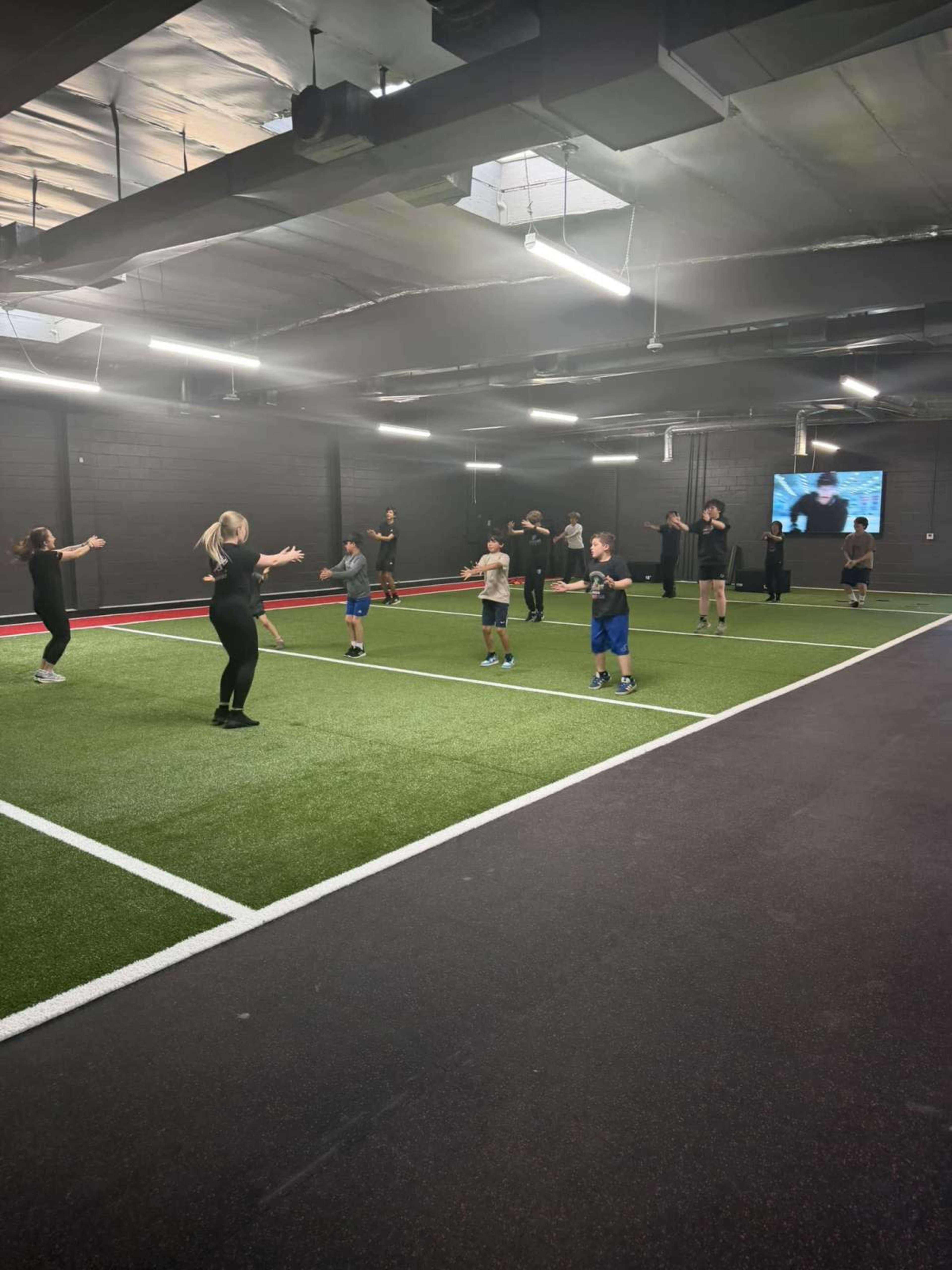 A group of children and adults are participating in a workout session inside a gym with artificial turf and neon lights.