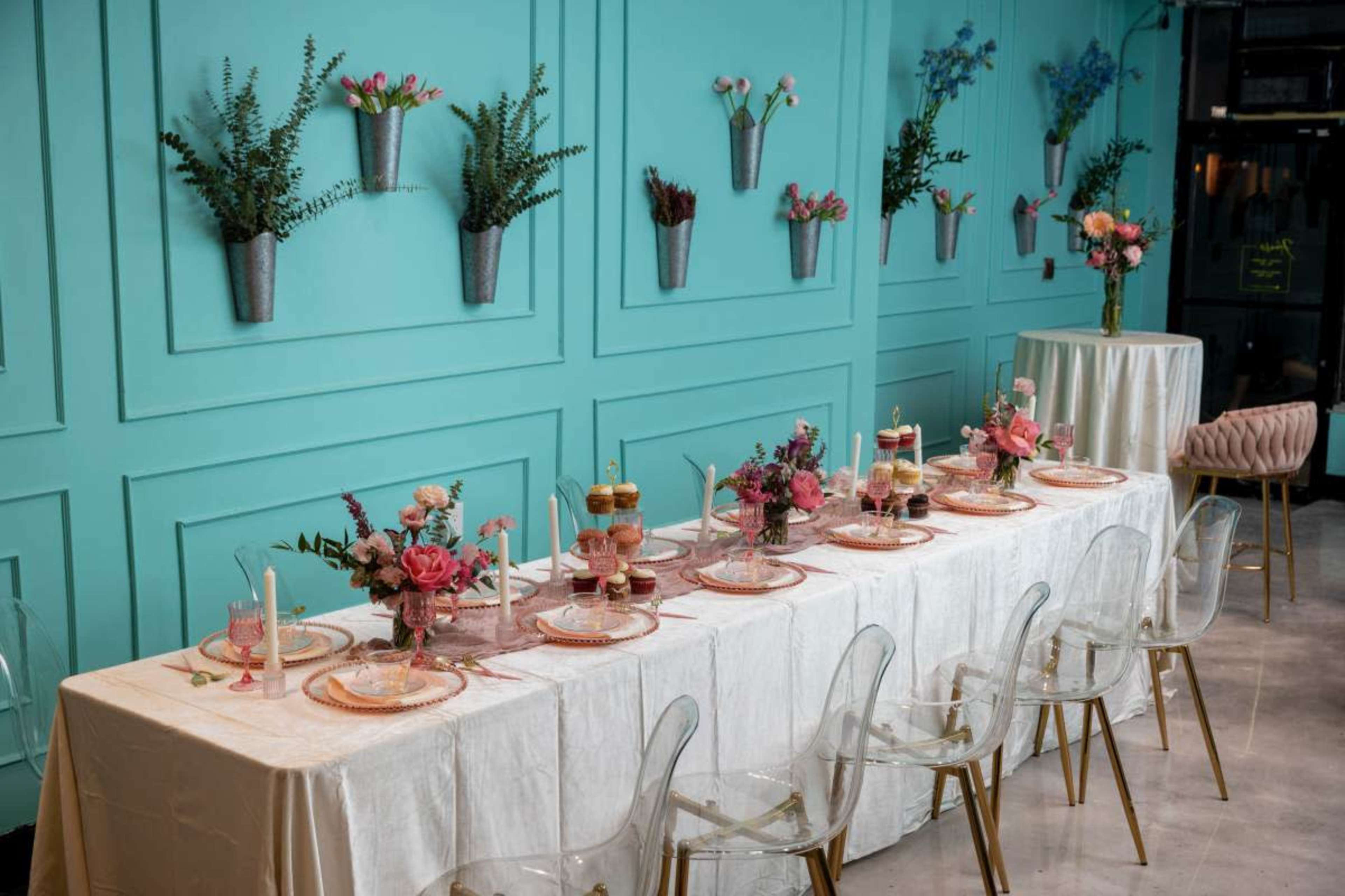 A long dining table is elegantly set with floral arrangements, tableware, and decorative elements against a turquoise wall adorned with potted plants.