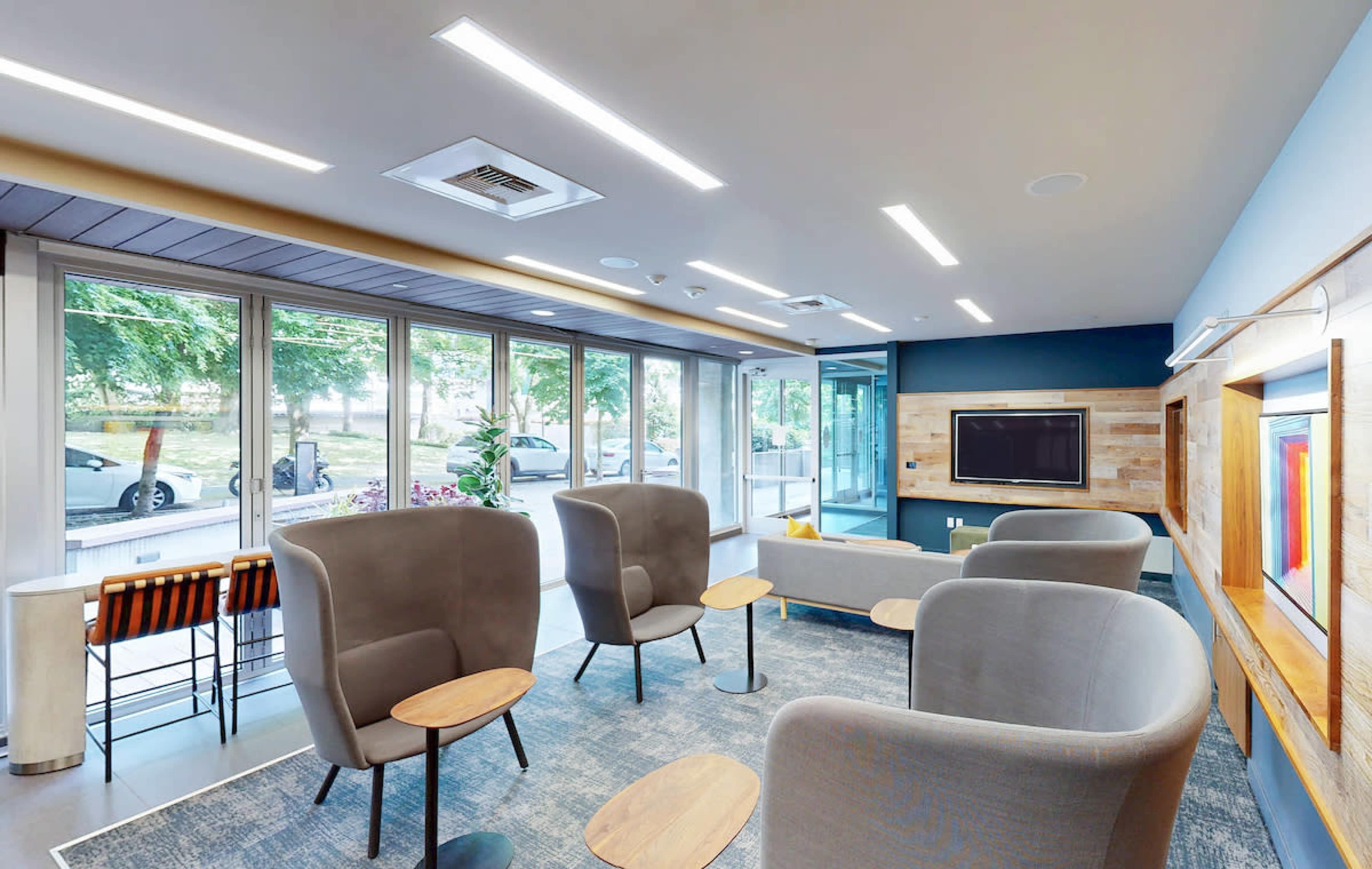 The image shows a modern lounge area with comfortable seating, a television, and large windows that provide natural light.