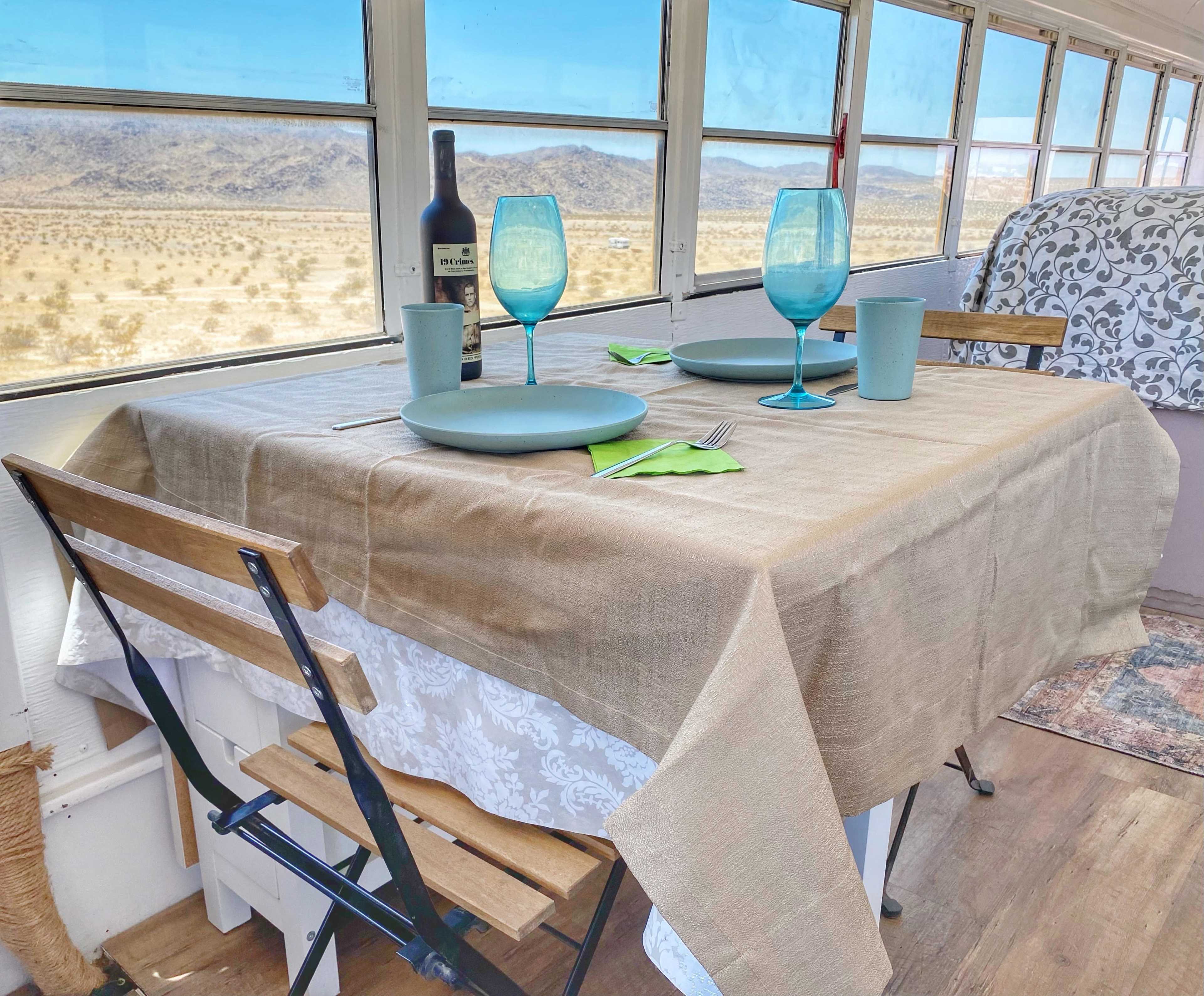A set dining table with blue glasses, plates, and utensils is positioned in front of a large window overlooking a desert landscape.