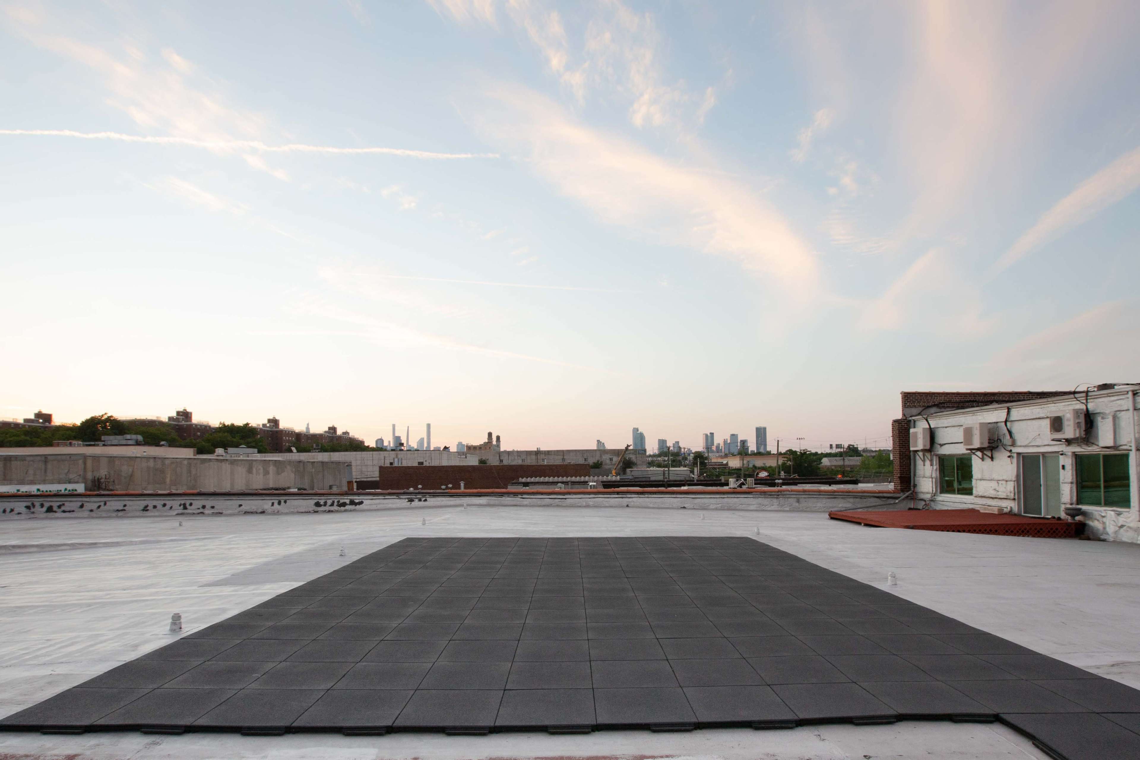 A flat rooftop with a square, dark tile area overlooks a city skyline under a pastel sky.
