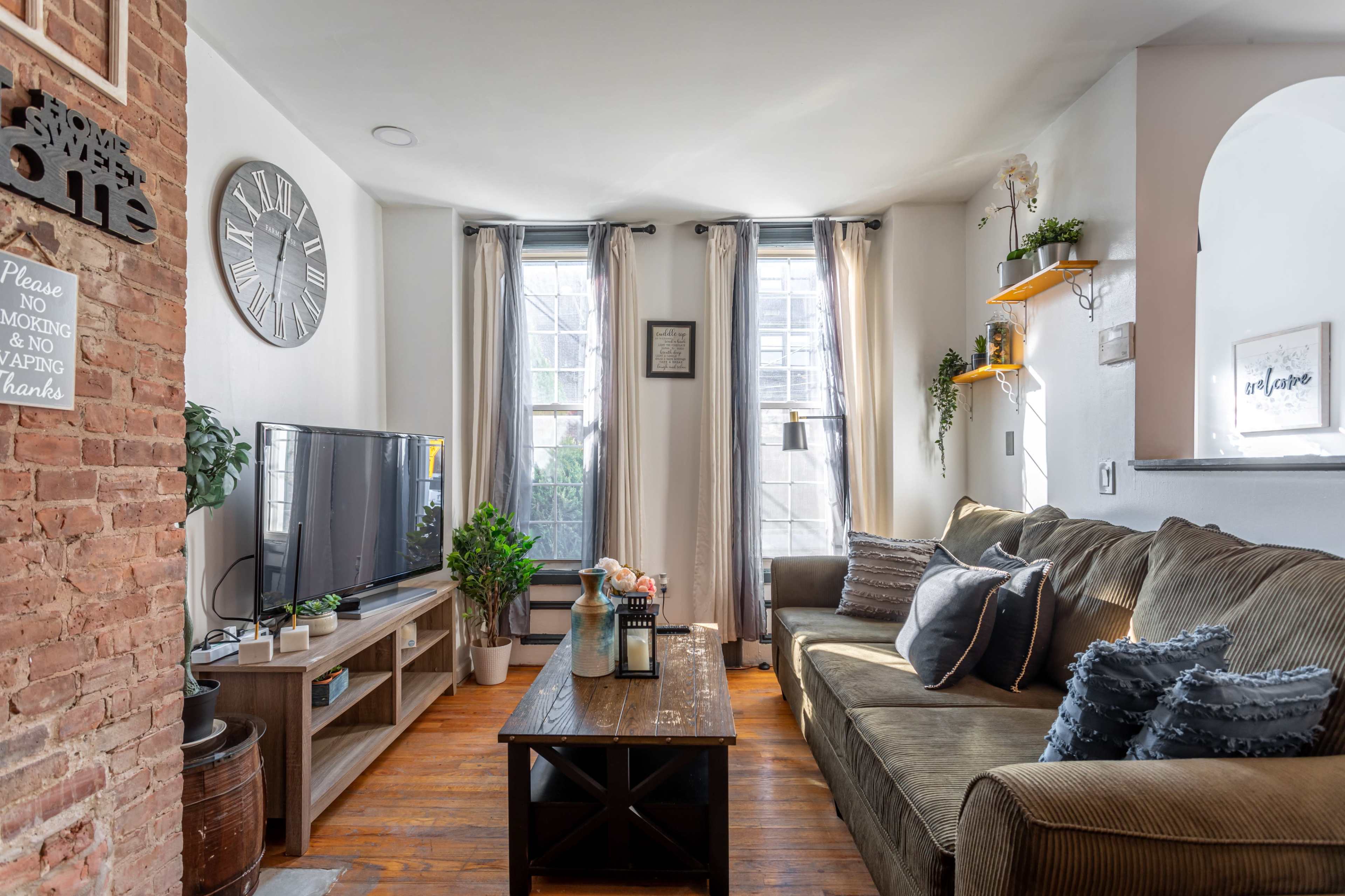 A cozy living room with a large sofa, a television, and plants, featuring natural light coming through two windows.