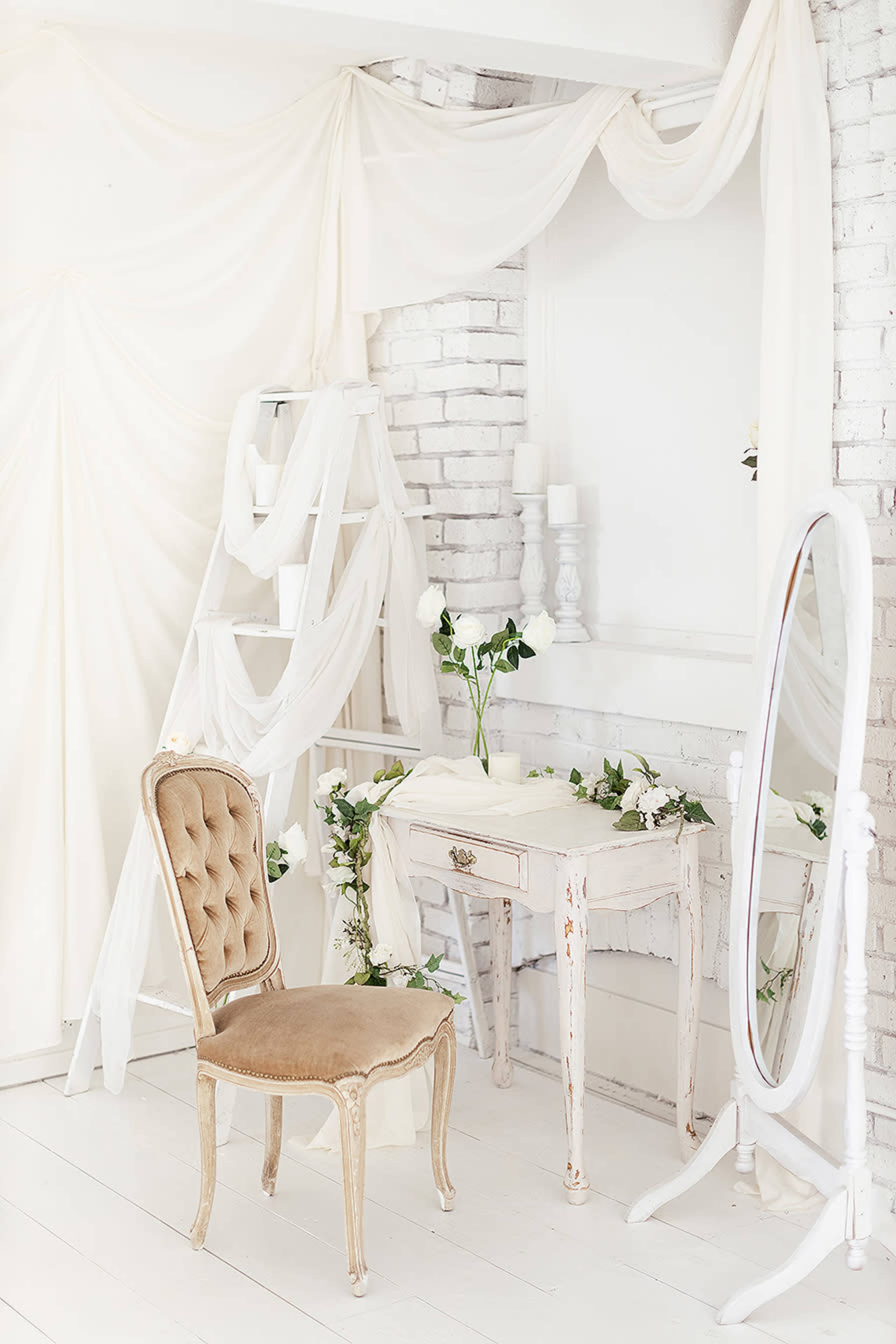 The scene features a light-filled room with a vintage chair, a small white table, a full-length mirror, and decorative drapery and greenery against a brick wall.