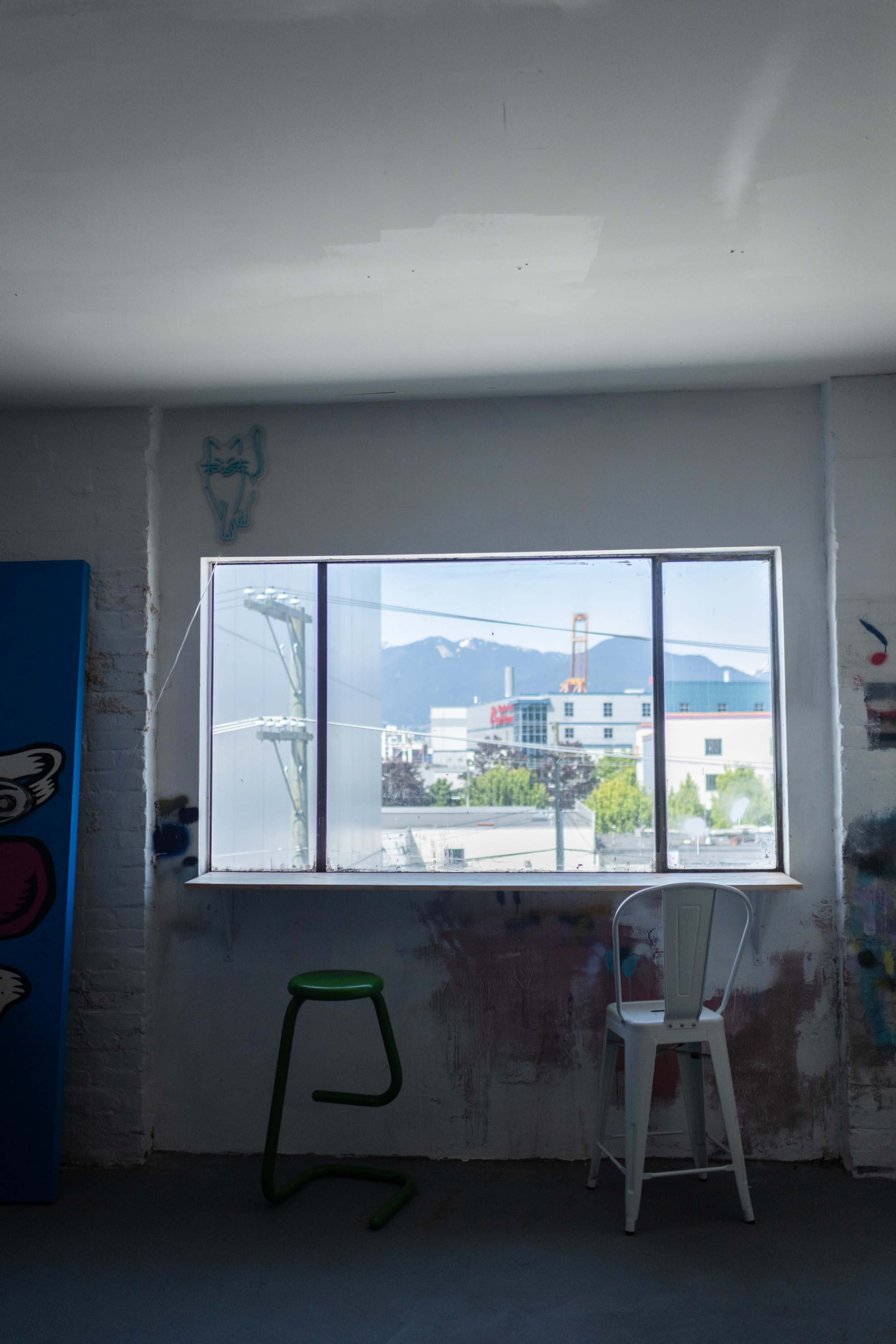 The image shows a bright room with a large window revealing a view of mountains, a utility pole, and buildings outside, along with a green stool and a white chair inside.