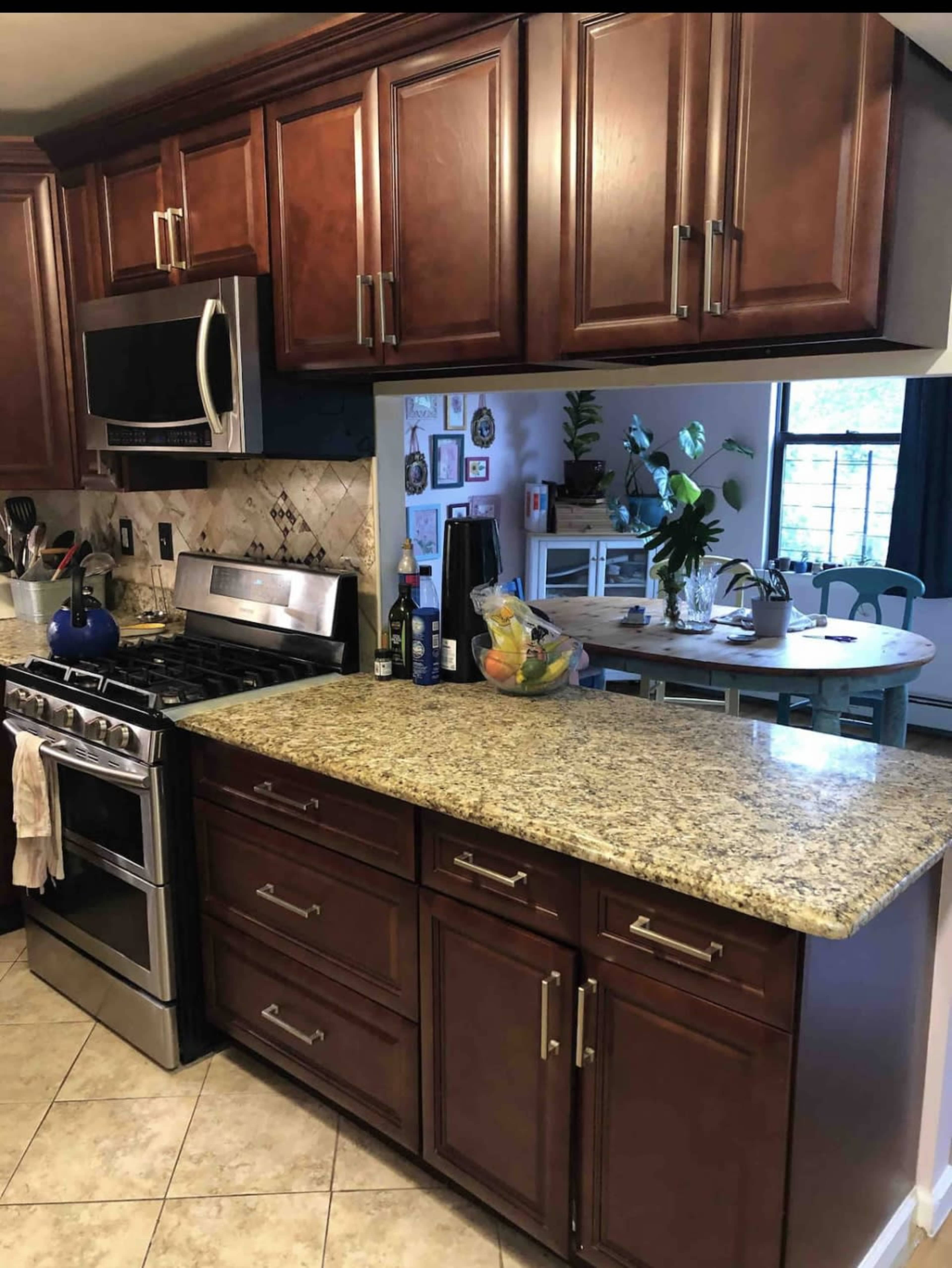 A kitchen with dark wood cabinetry, a stainless steel stove and microwave, and a granite countertop, with a dining area visible in the background.