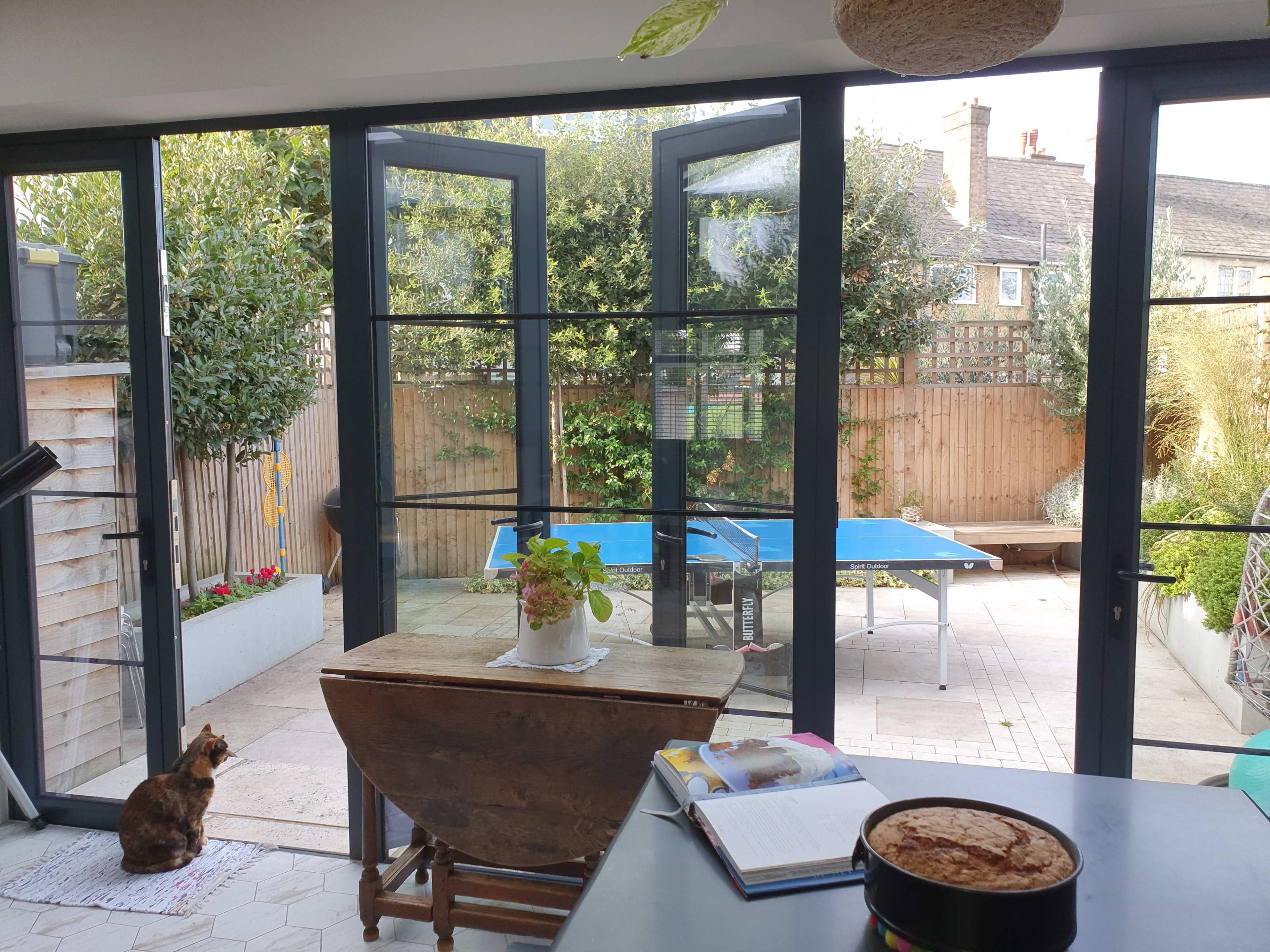 The image shows a bright interior space with large glass doors open to a garden, featuring a ping pong table, a wooden table, and a cat sitting on the floor.