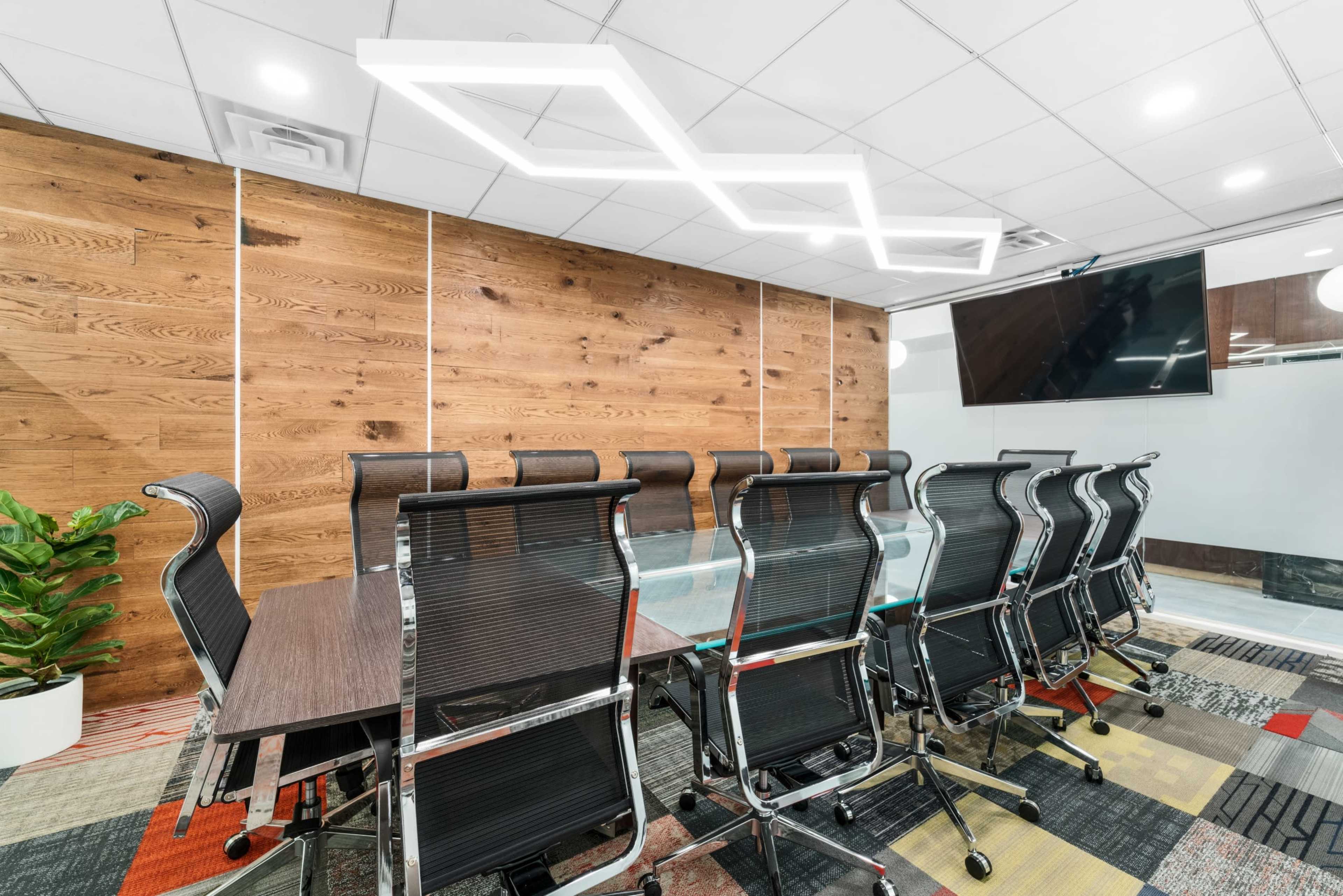 A modern conference room features a long table surrounded by black ergonomic chairs, with a wooden accent wall and contemporary lighting above.
