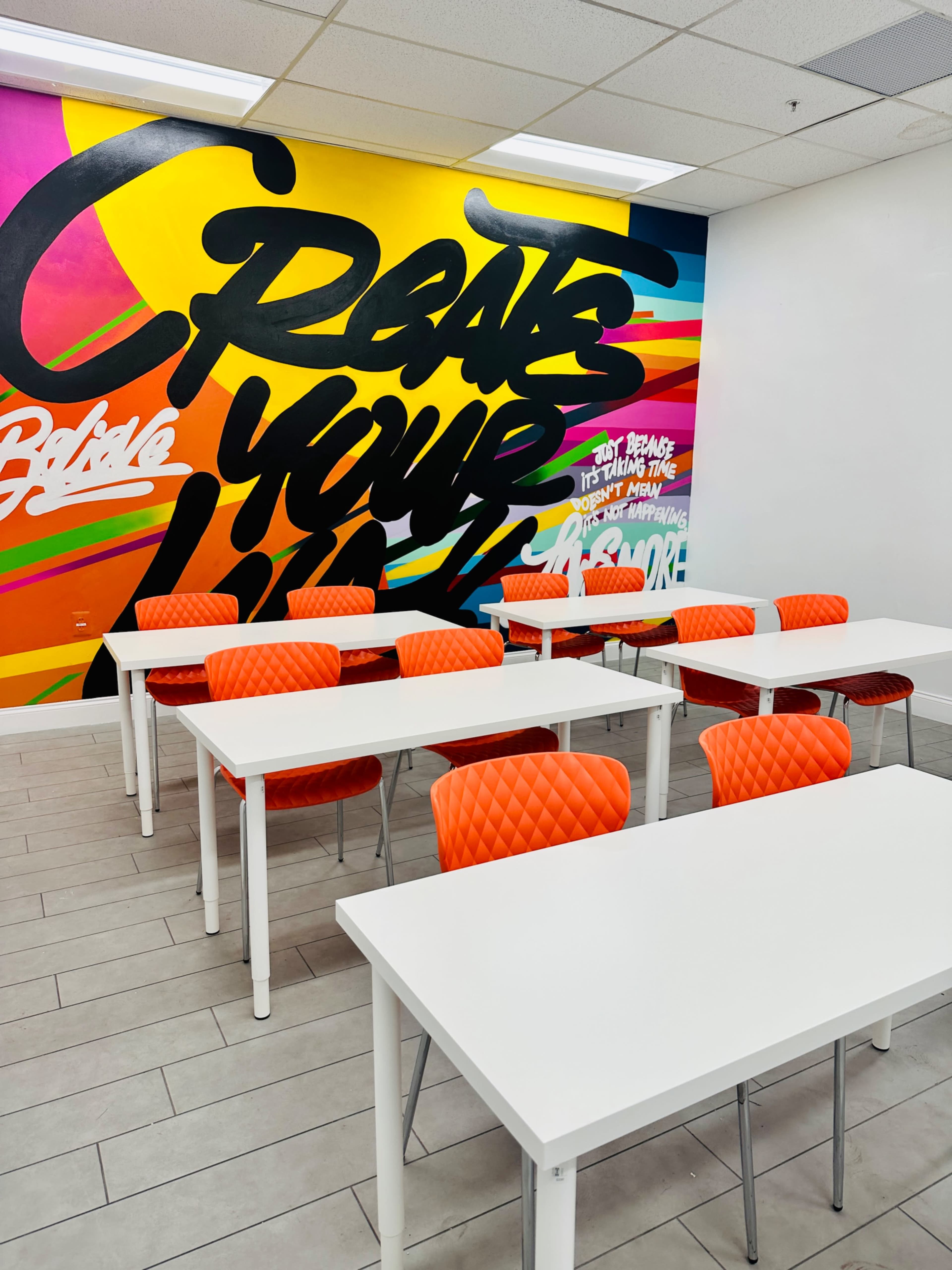 A brightly colored classroom features white tables arranged in rows with orange quilted chairs and a vibrant mural on the wall.