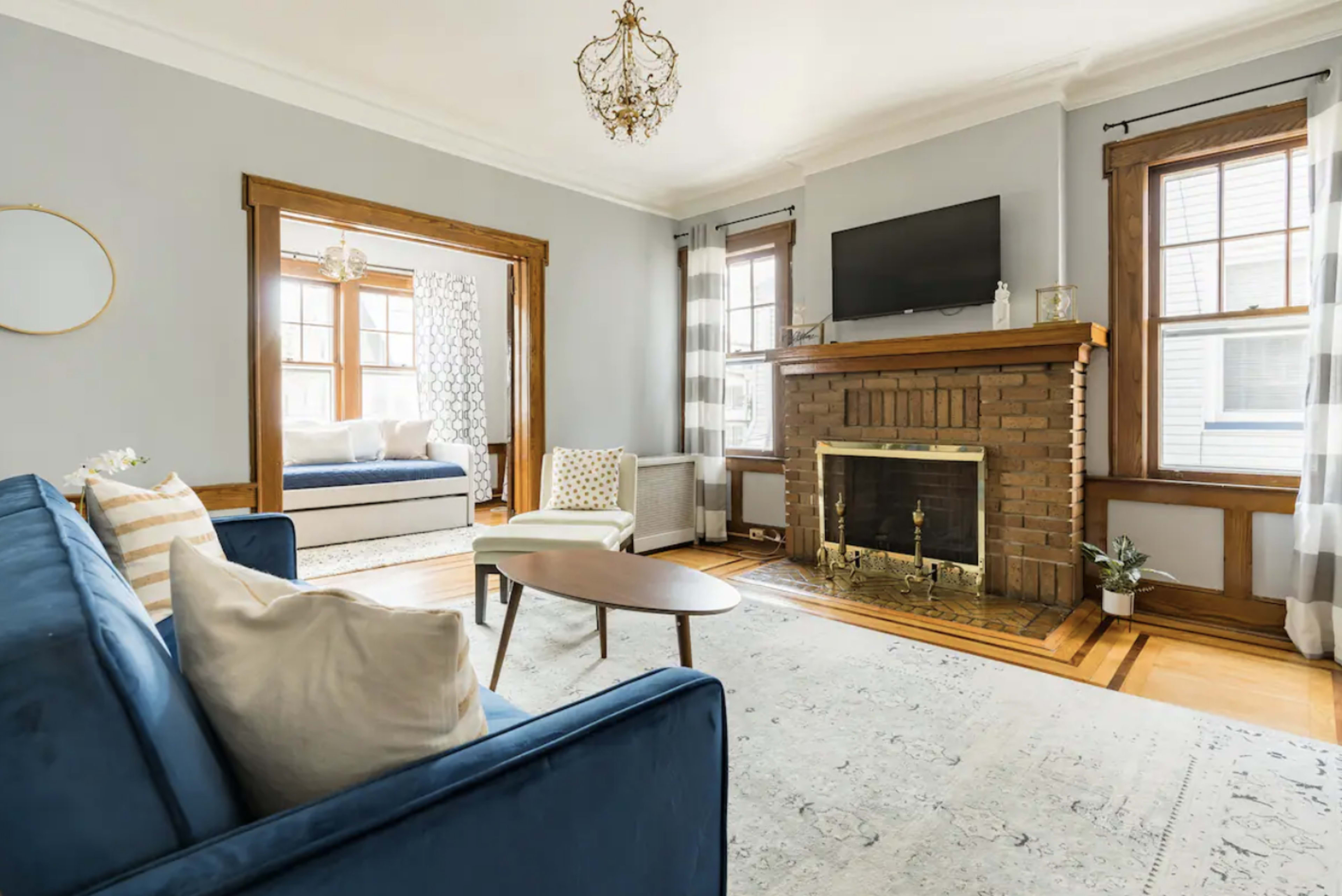 The living room features a blue sofa, a coffee table, and a brick fireplace, with large windows allowing natural light and an adjoining room visible in the background.