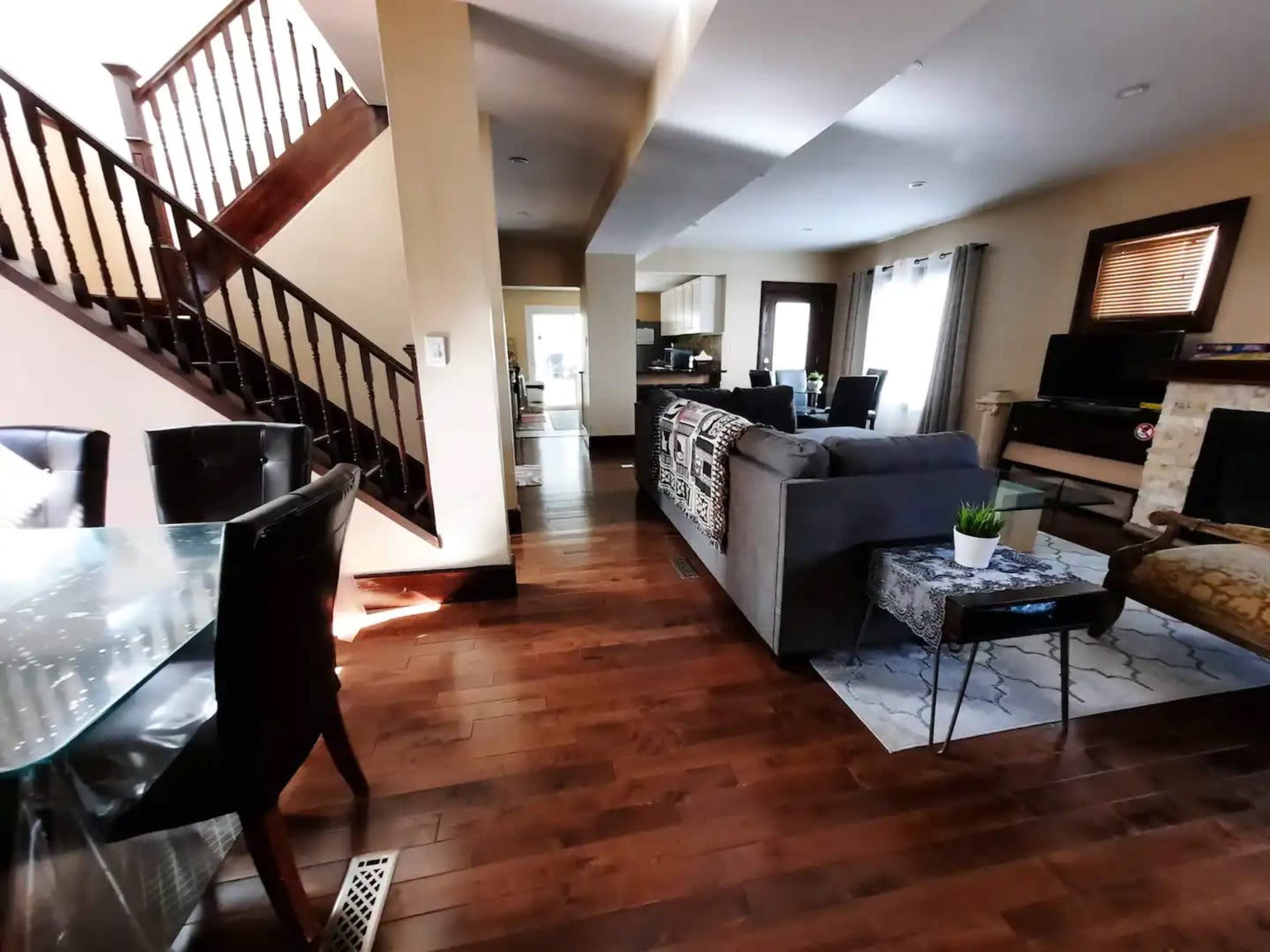 A living room area with hardwood floors, a staircase in the background, and a dining area with glass table and black chairs.