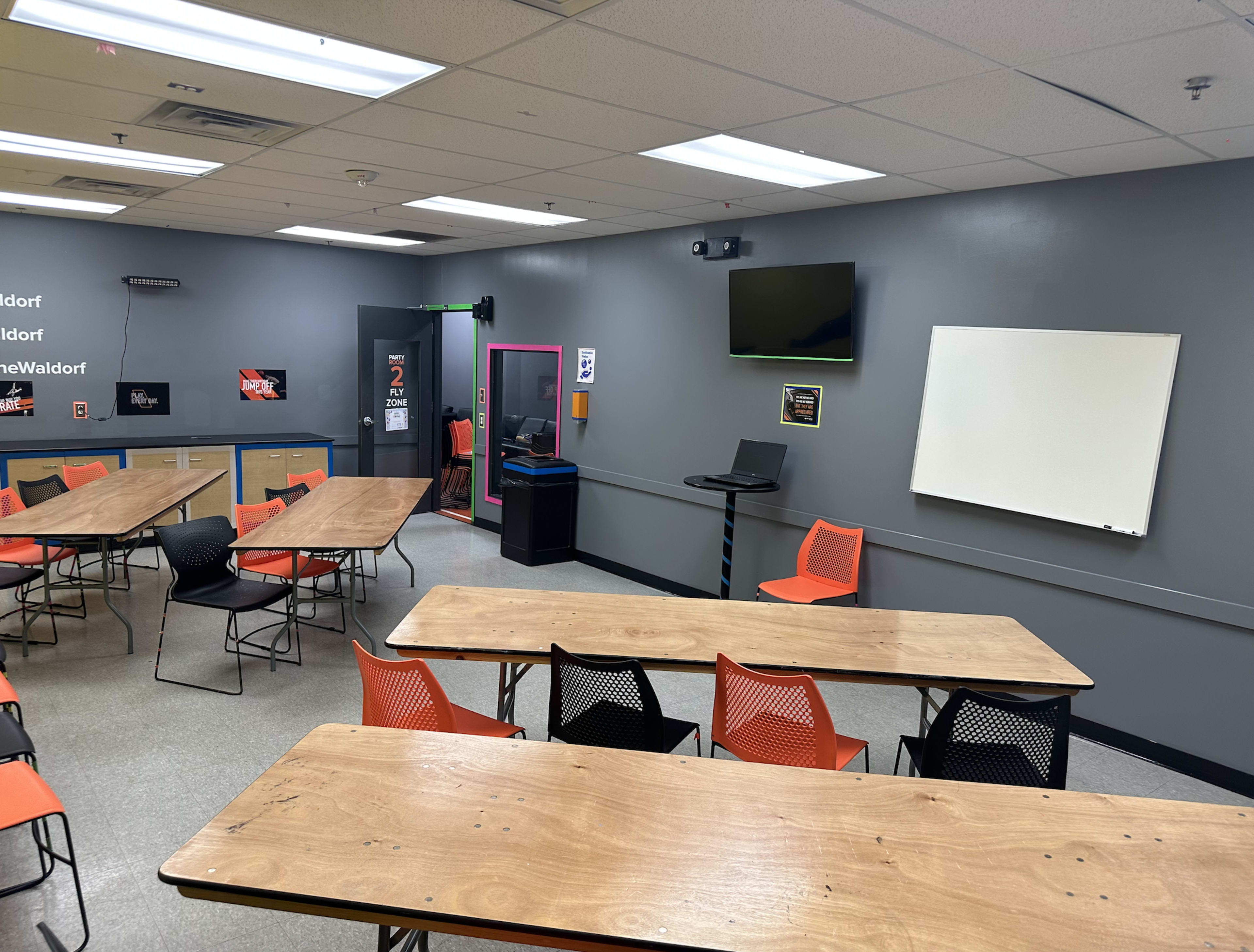 A classroom is equipped with tables, chairs, a television, a whiteboard, and a computer station against a gray wall.