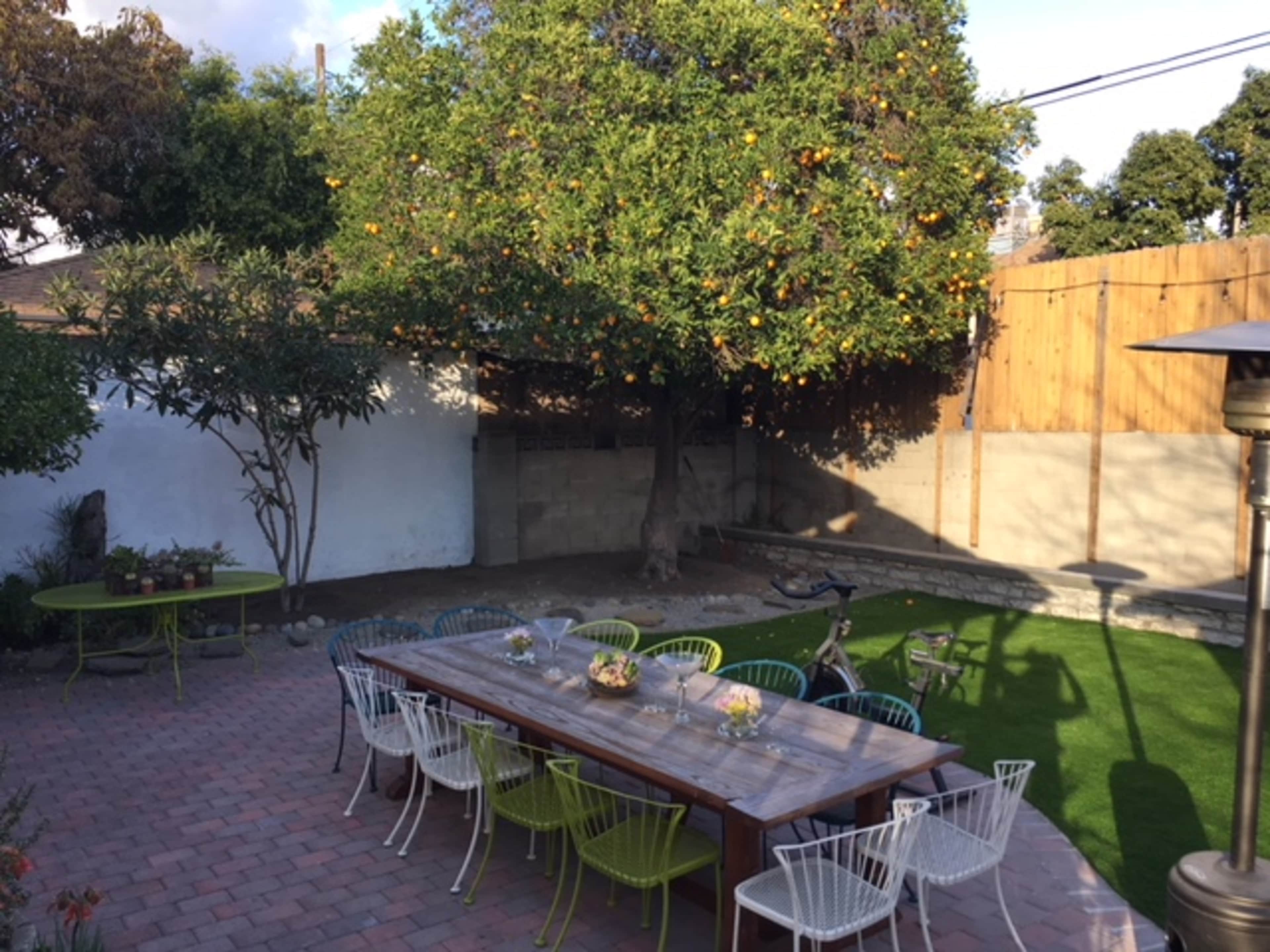 The image shows a landscaped backyard with a long wooden table surrounded by chairs, a large fruit-bearing tree, and a well-maintained lawn.