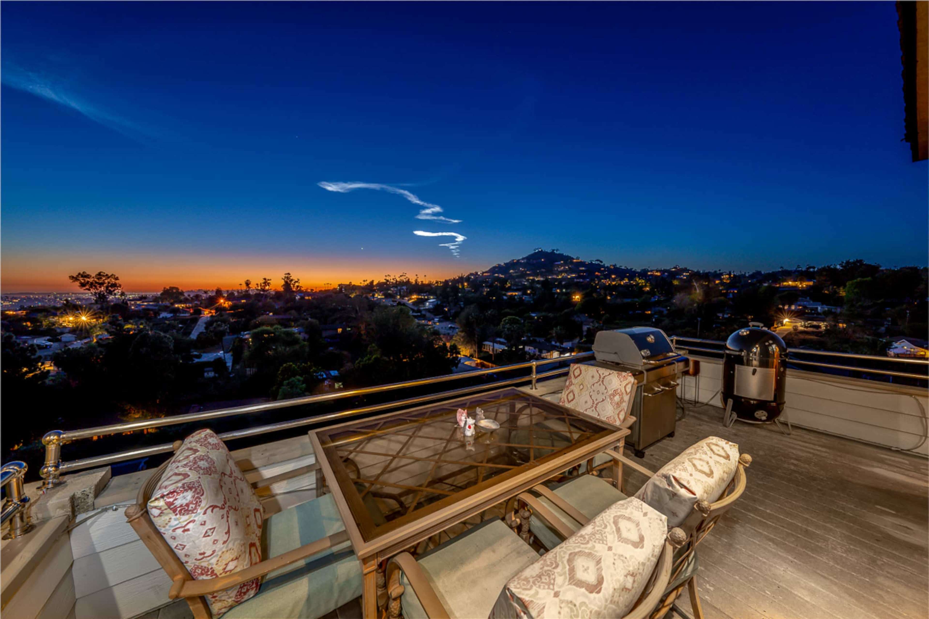 A balcony features a wooden table and chairs overlooking a cityscape at sunset, with a grill nearby and streaks of cloud in the sky.