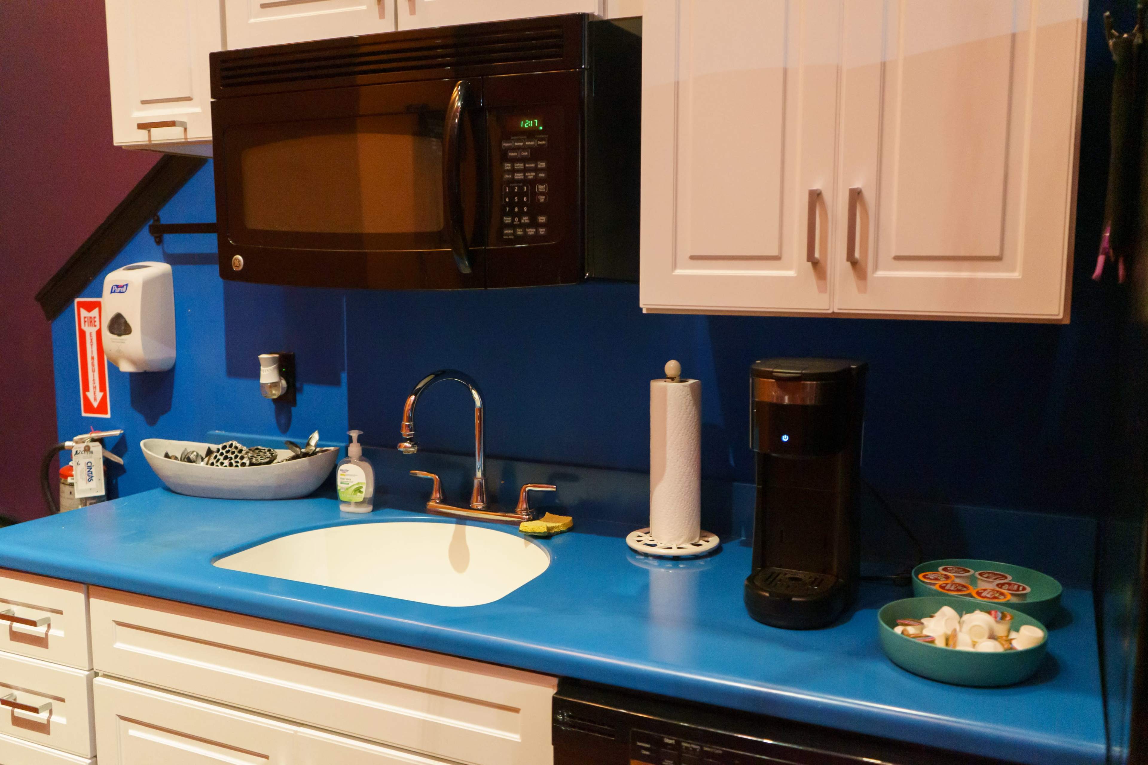 A kitchen countertop with a microwave, a coffee maker, a sink, hand soap, and colorful dishware on a bright blue surface.