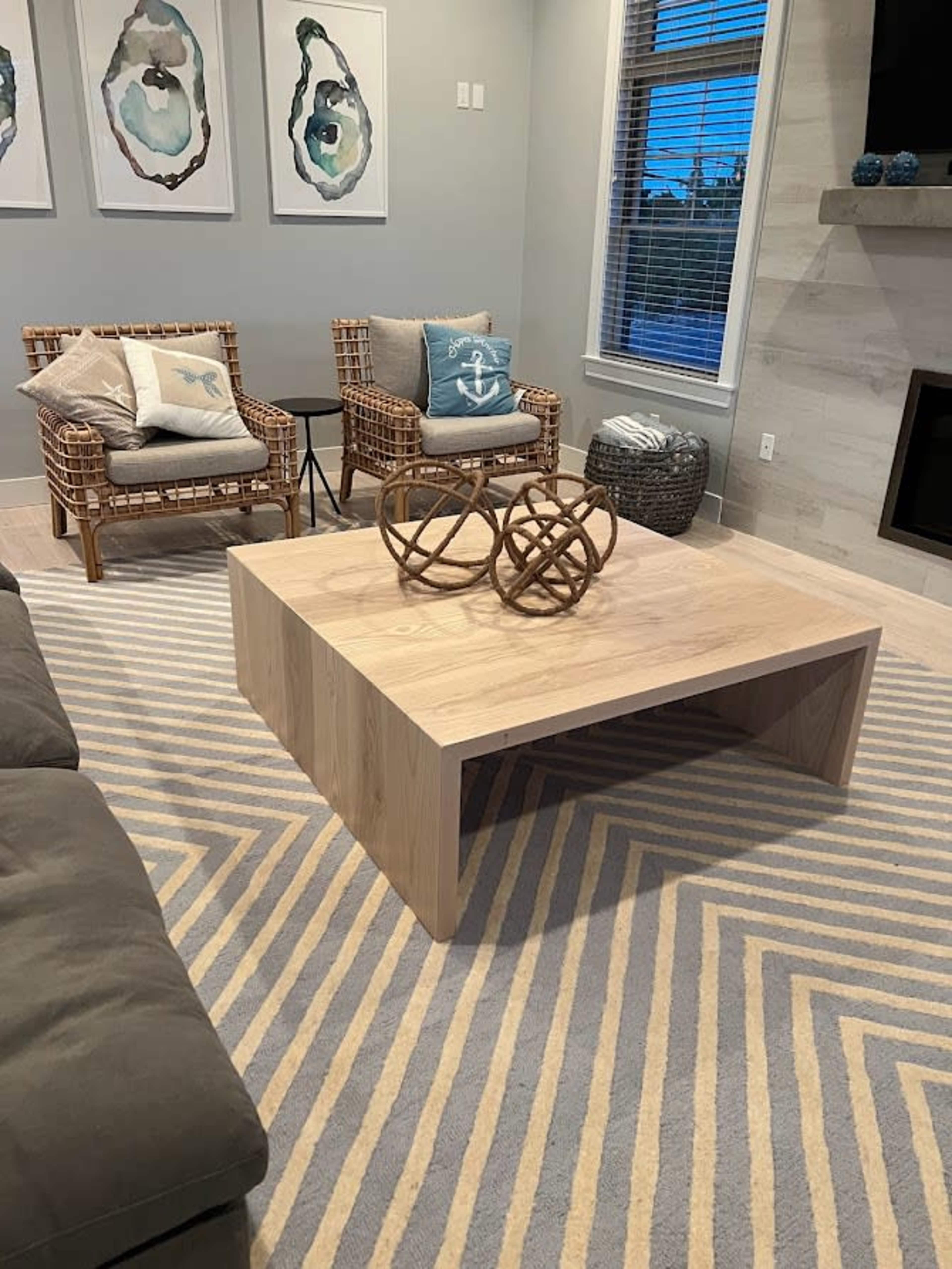 The image shows a living room with a light wooden coffee table, two wicker chairs, and a patterned rug beneath them.