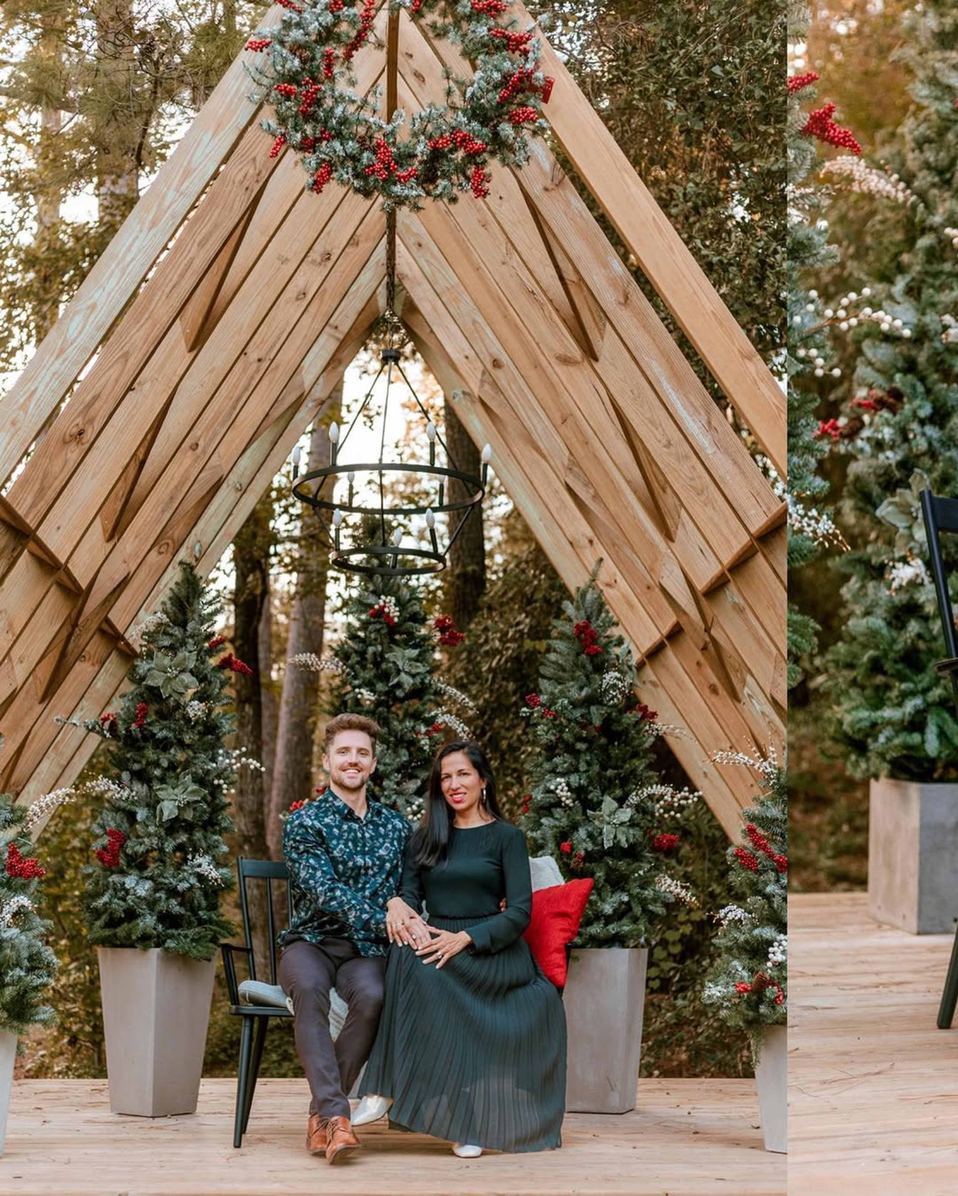 A man and a woman sit together on a chair in front of decorated Christmas trees and a wooden arch adorned with a wreath.