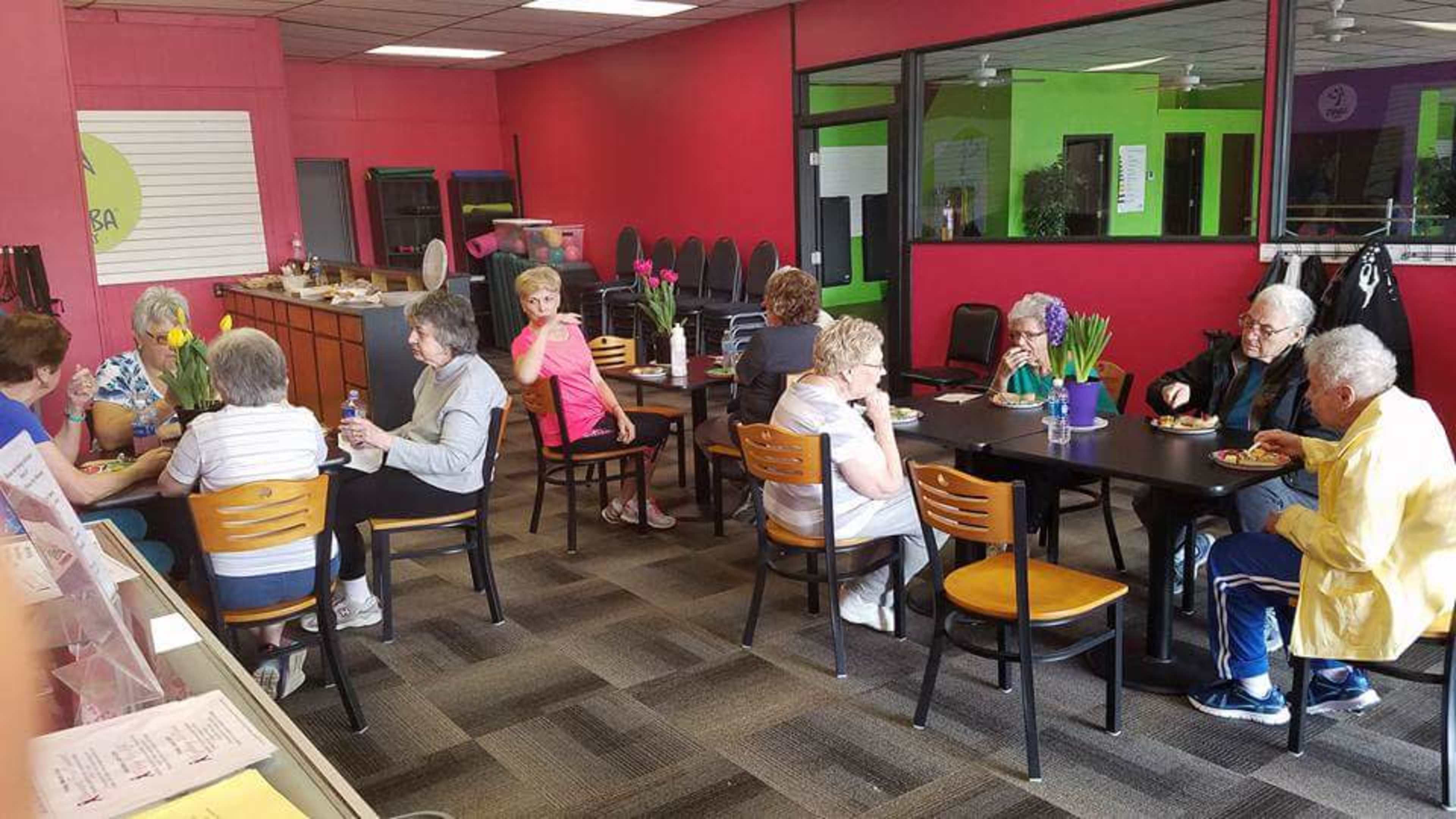 A group of elderly individuals socialize and enjoy refreshments around tables in a brightly colored community center.
