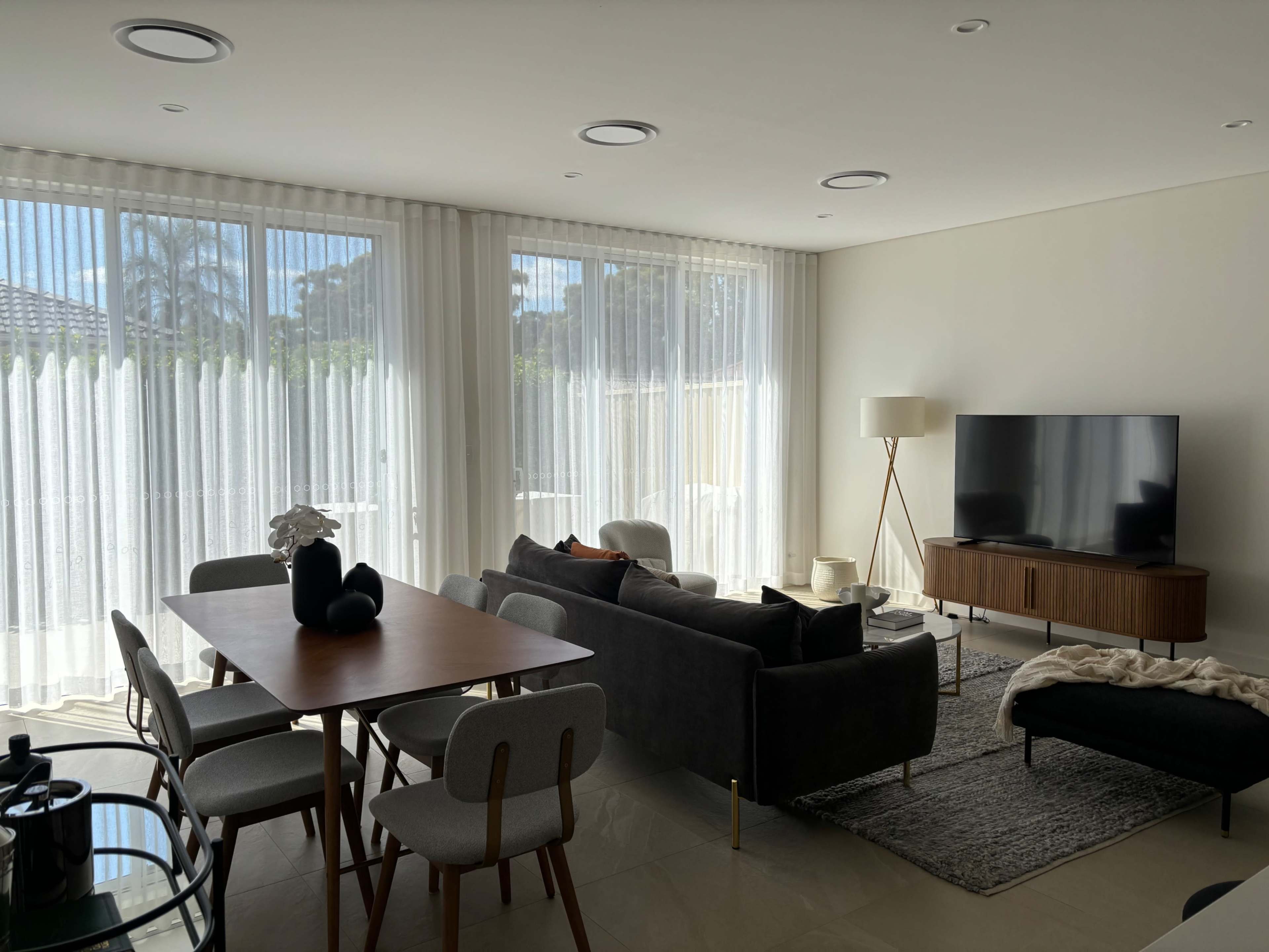The image shows a modern living room with large windows covered by sheer curtains, a dining table, a sofa, and a television on a low cabinet.
