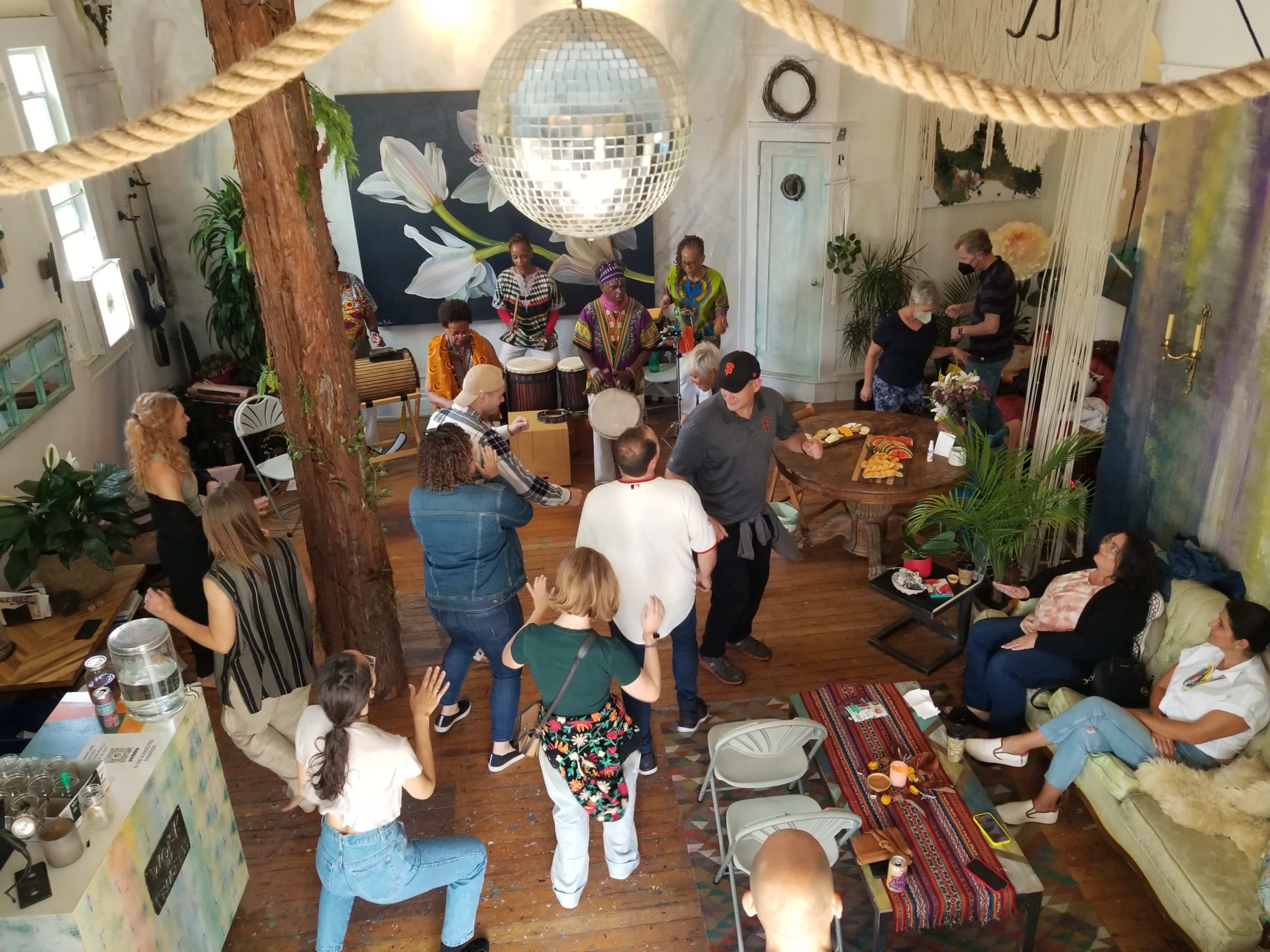 A diverse group of people gathers in a spacious room adorned with plants and artwork, engaging in a communal drumming session.