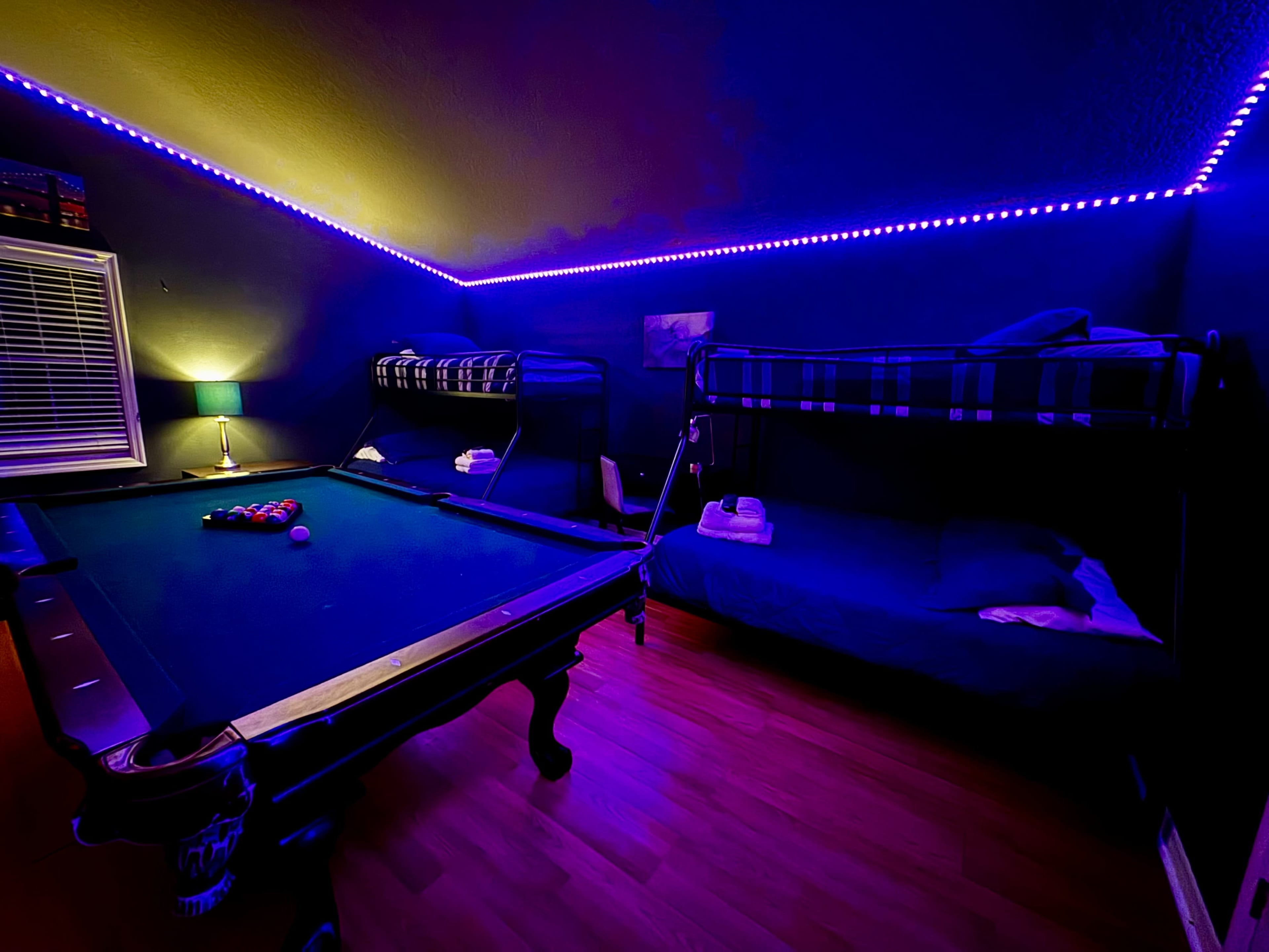 The image shows a dimly lit room featuring a pool table surrounded by bunk beds, with LED lights illuminating the ceiling.