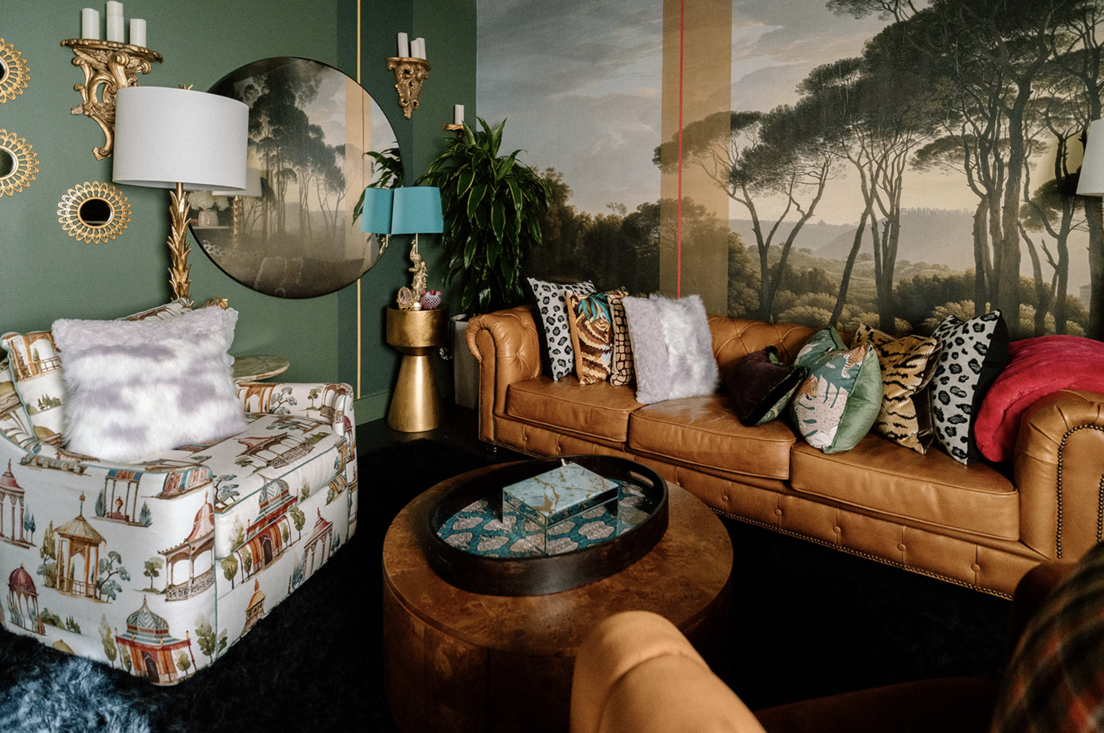 A stylish living room featuring a mix of patterned chairs, a leather couch, and decorative accessories against a mural of trees.