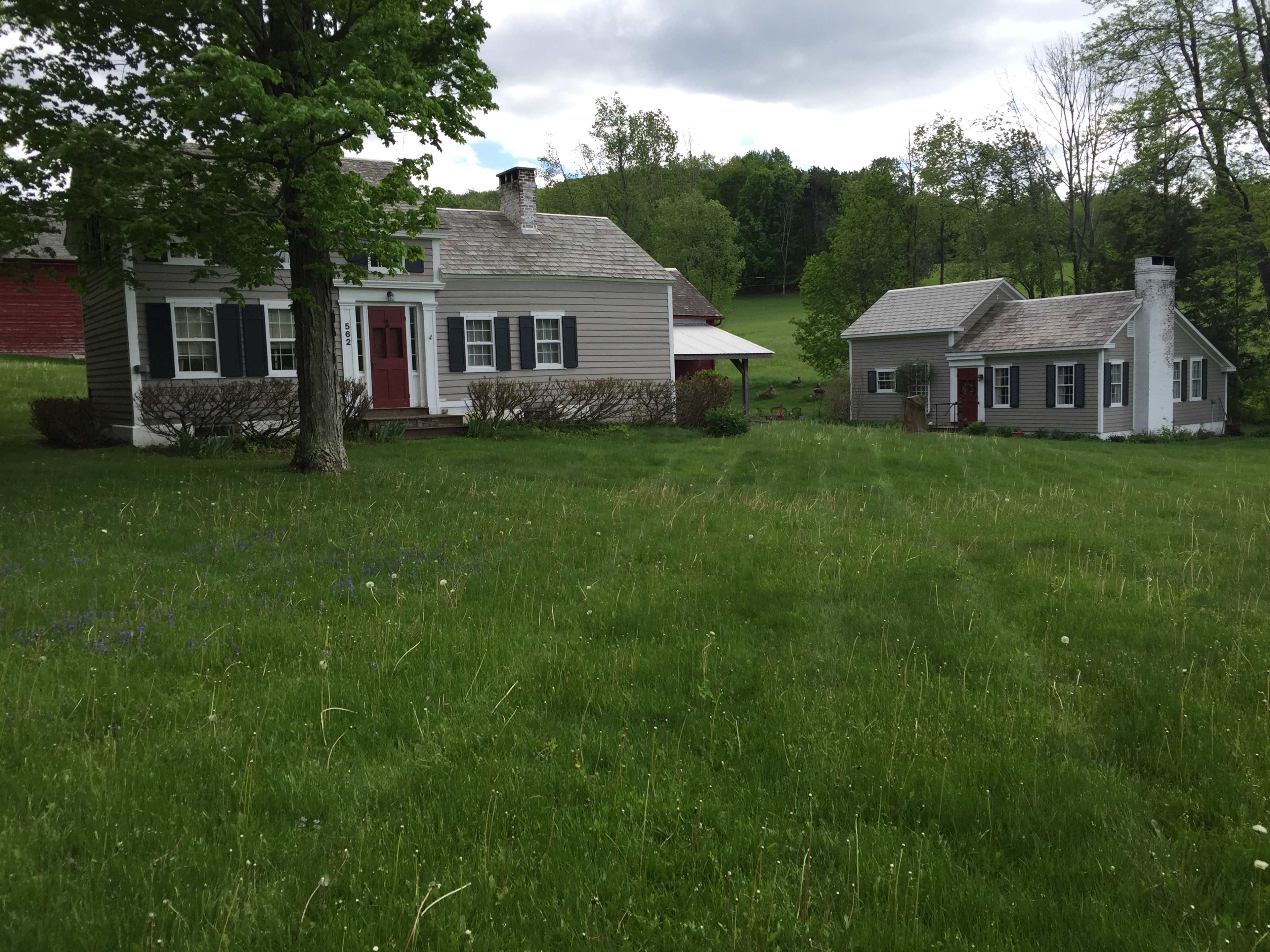 The image shows two gray houses with red doors set in a grassy area surrounded by trees.