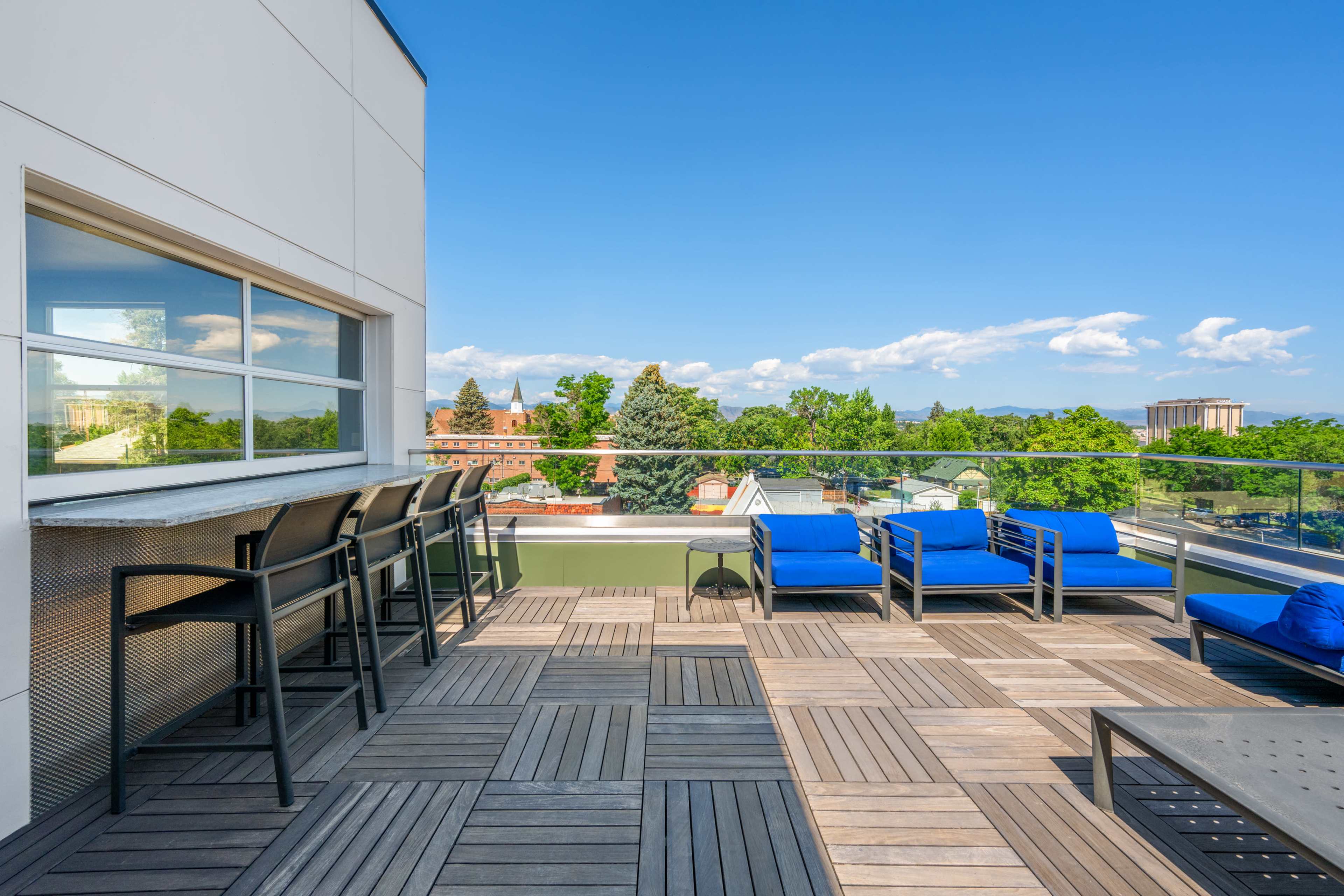 The image shows a rooftop terrace with blue chairs and a bar area, overlooking a green landscape and clear blue sky.