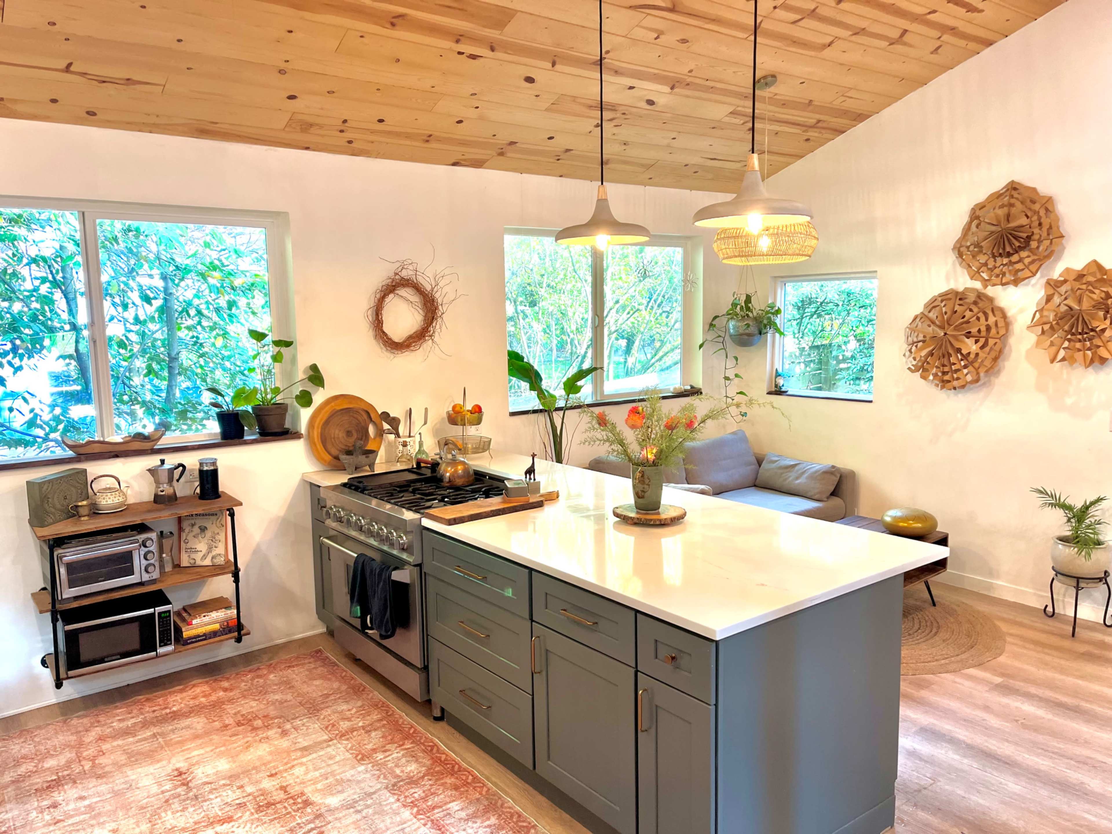 The image shows a modern kitchen with a central island, a stove, and several windows allowing natural light to illuminate the space, along with a cozy seating area and decorative wall art.
