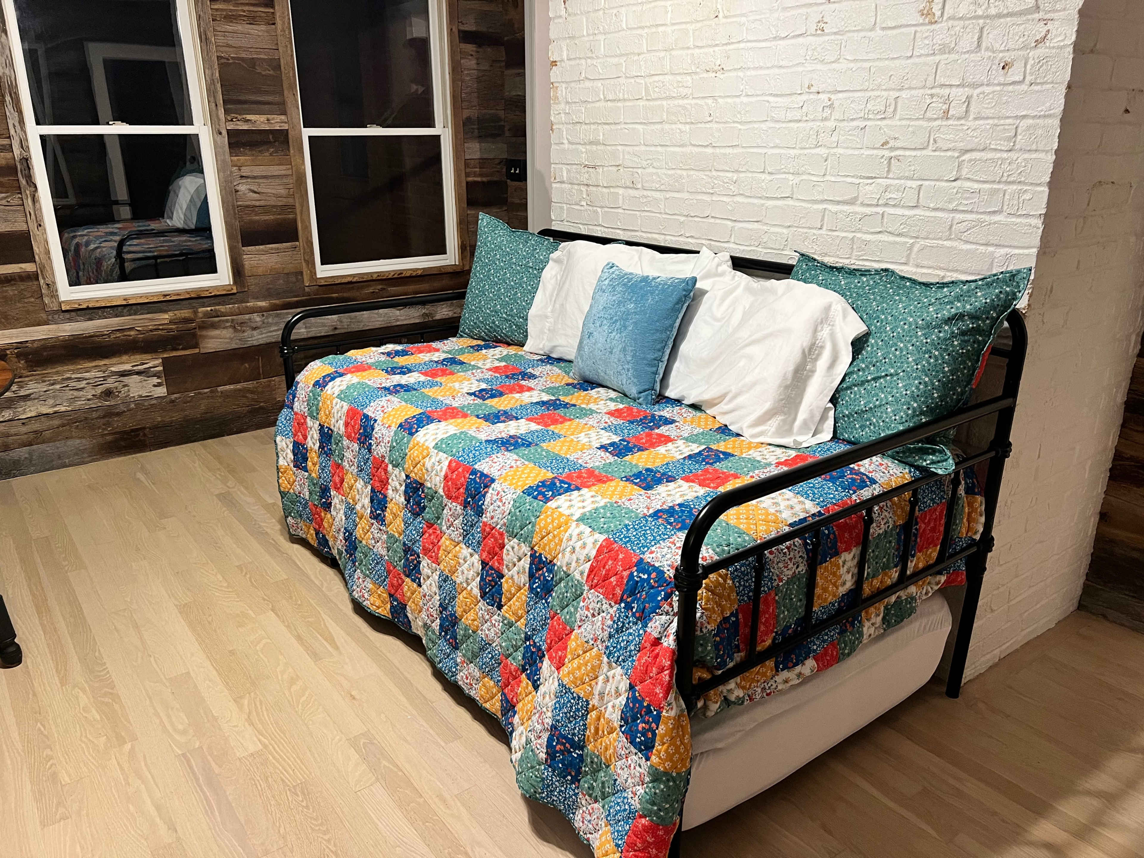 A daybed covered with a colorful quilt is positioned against a white brick wall in a room with wooden accents.