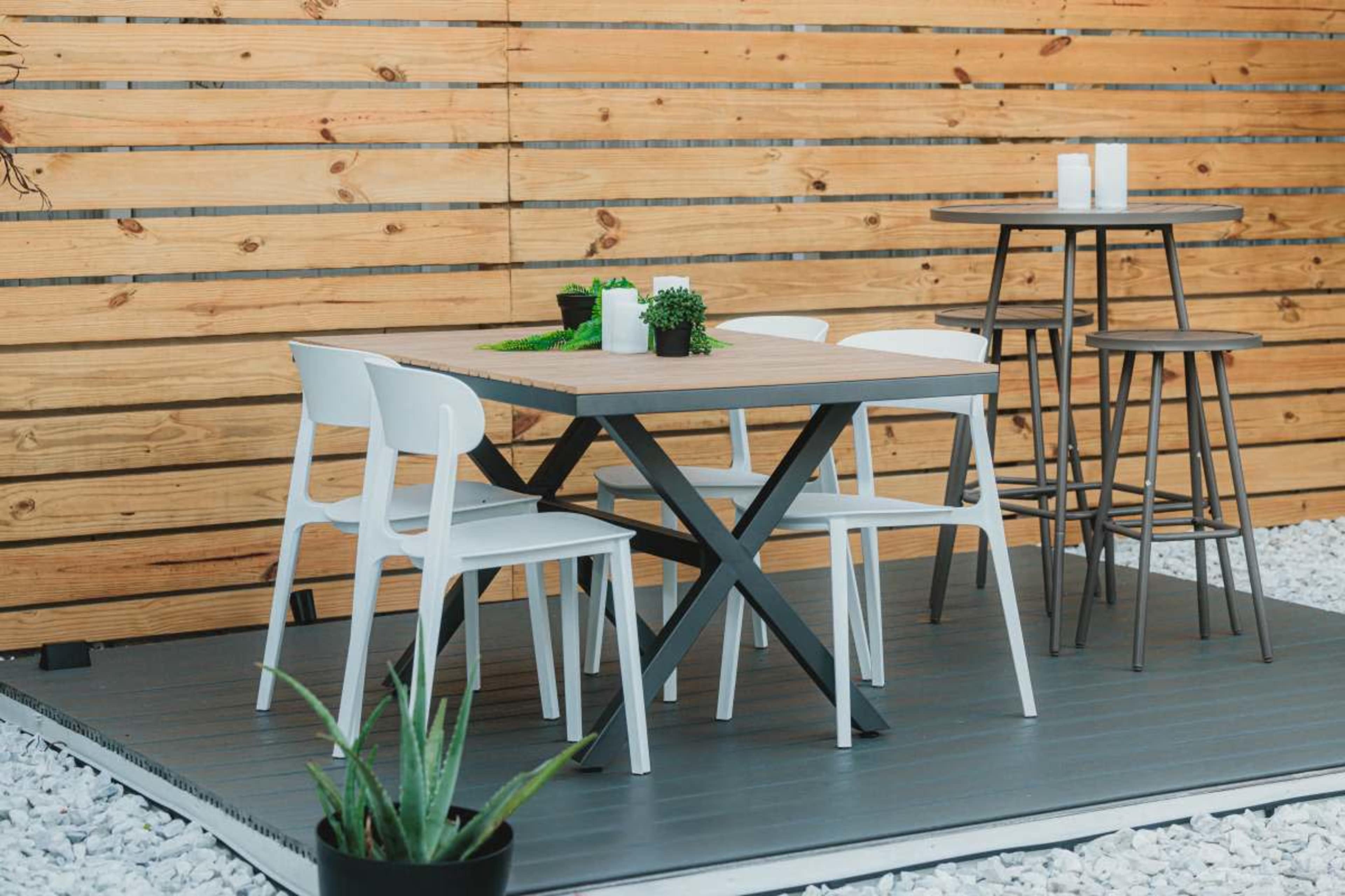 A wooden patio area features a table surrounded by white chairs, with a tall table and stools nearby, all set against a backdrop of wooden slats.