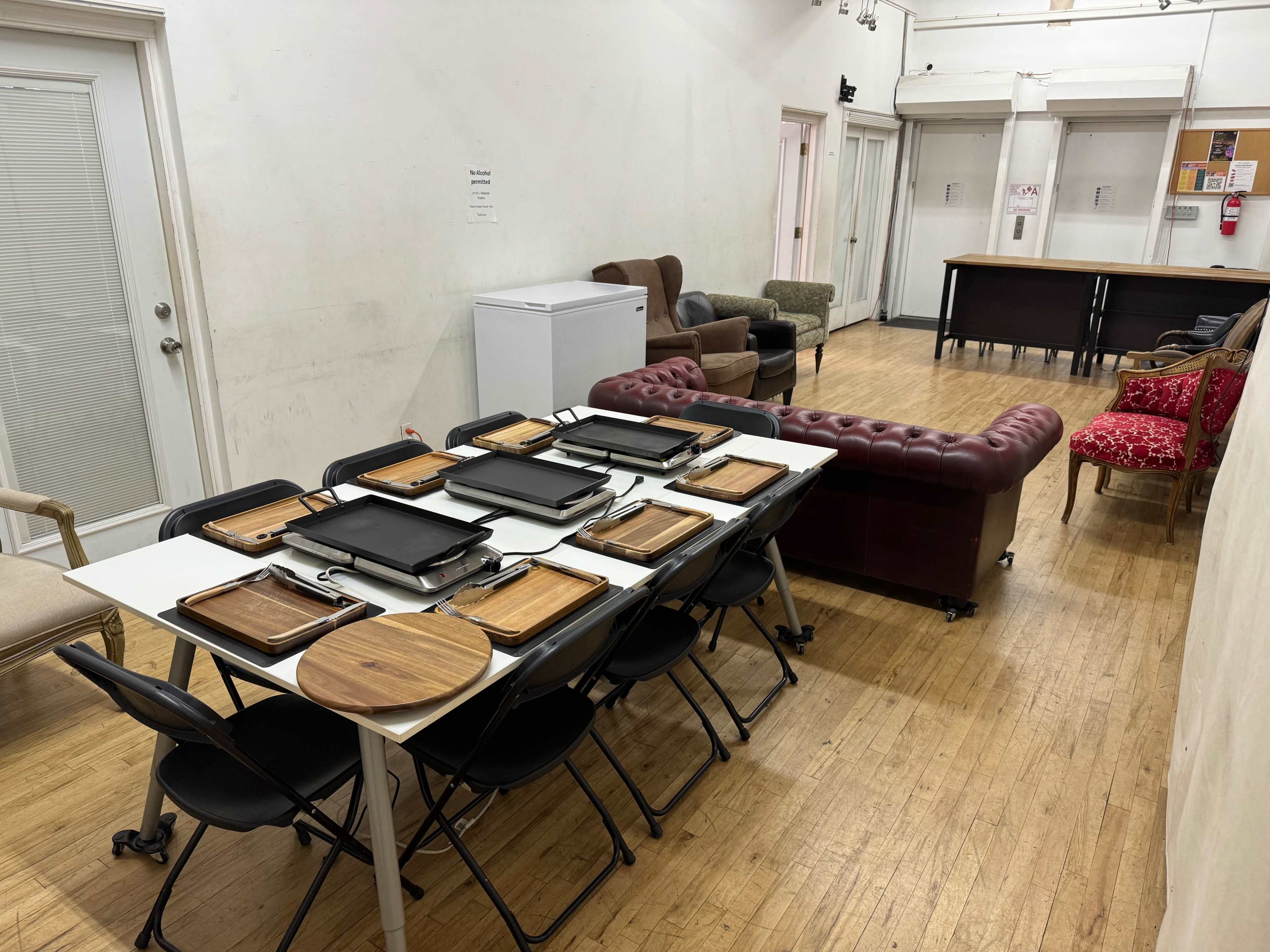 The image shows a large table with multiple trays set on it, surrounded by chairs, with a seating area featuring various couches in a hall-like space.
