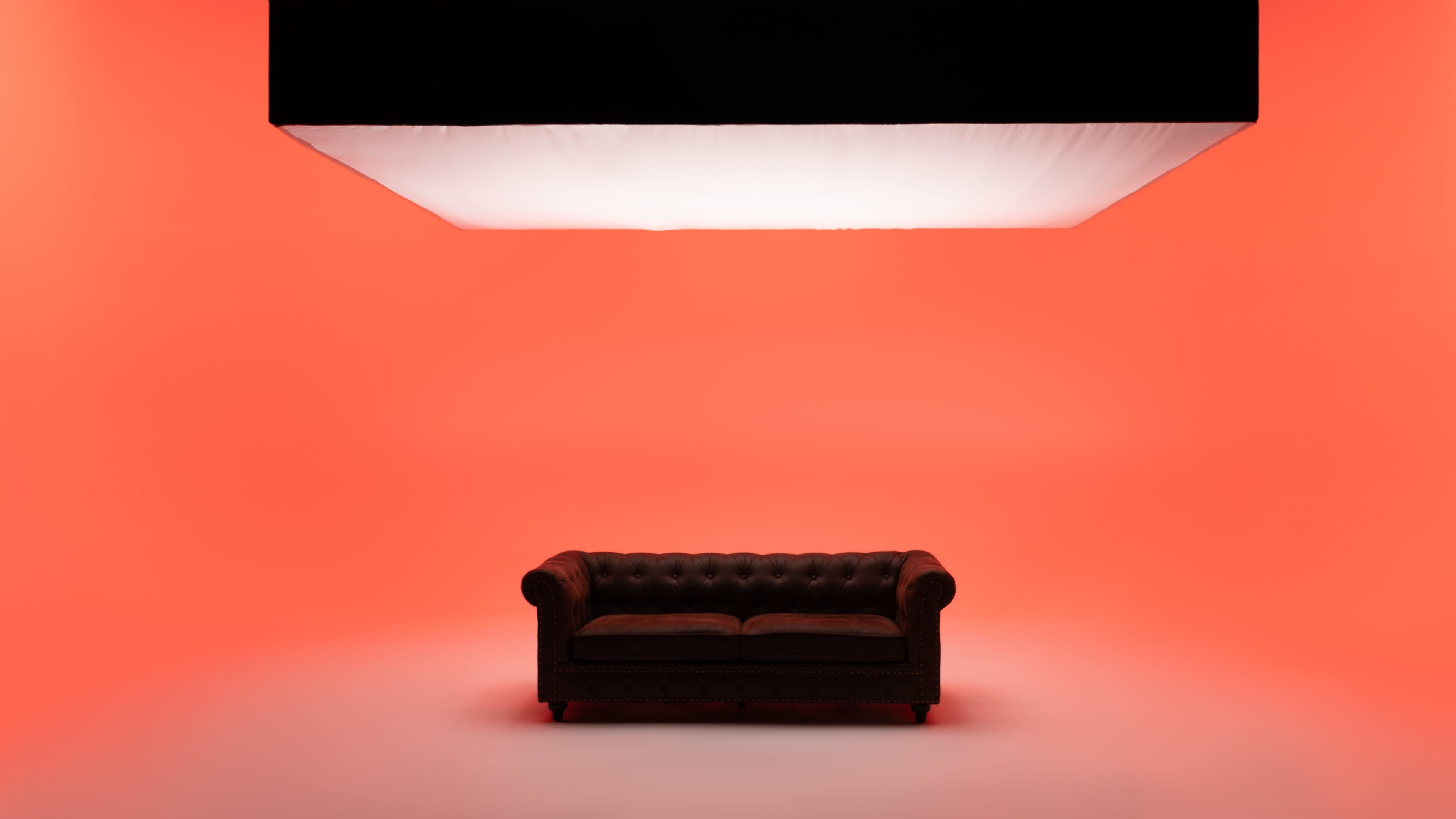 A dark leather chesterfield sofa is centered on a bright orange background beneath a large suspended light panel.
