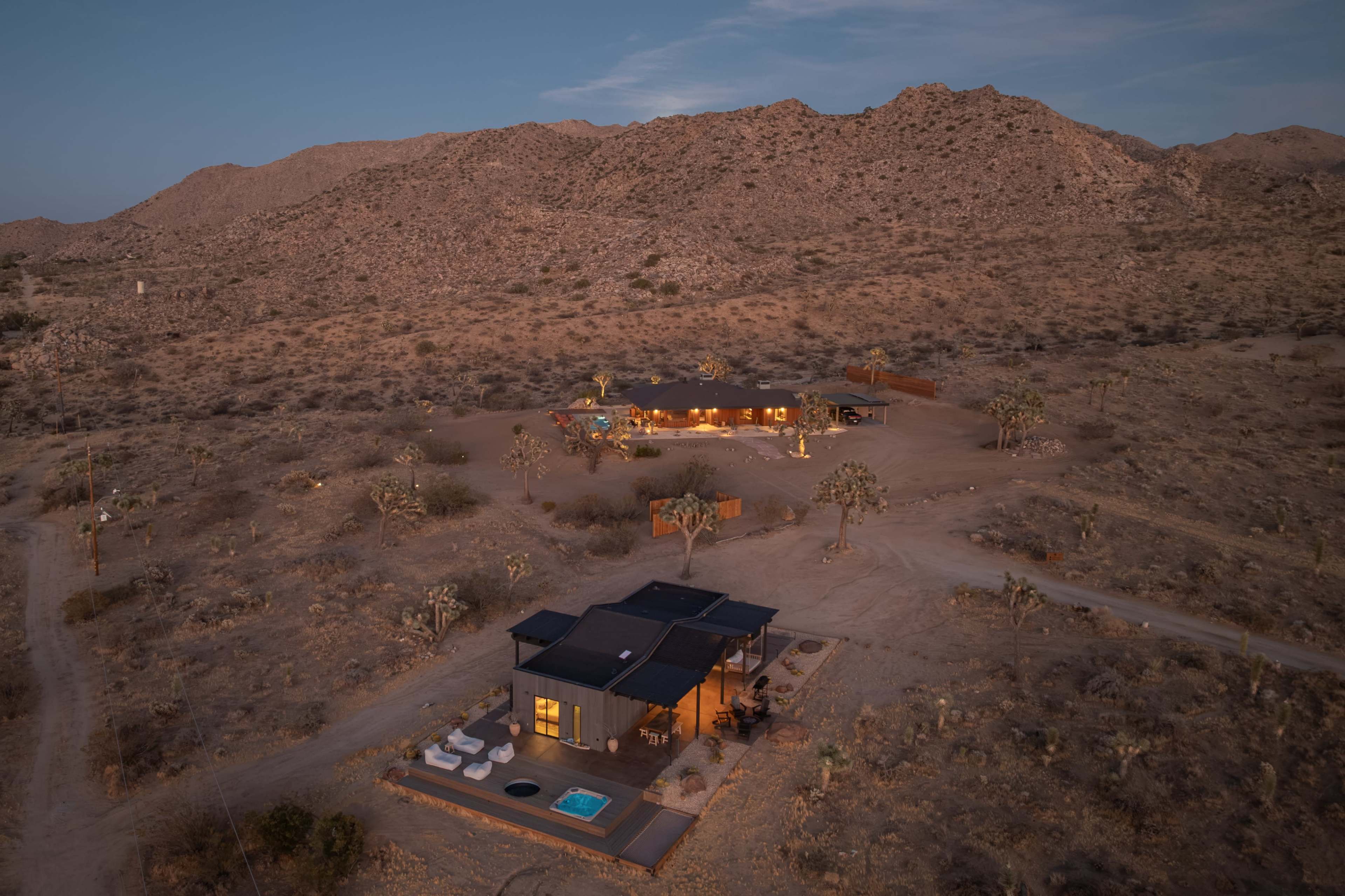 Aerial view of two modern houses situated in a desert landscape surrounded by mountains and sparse vegetation.