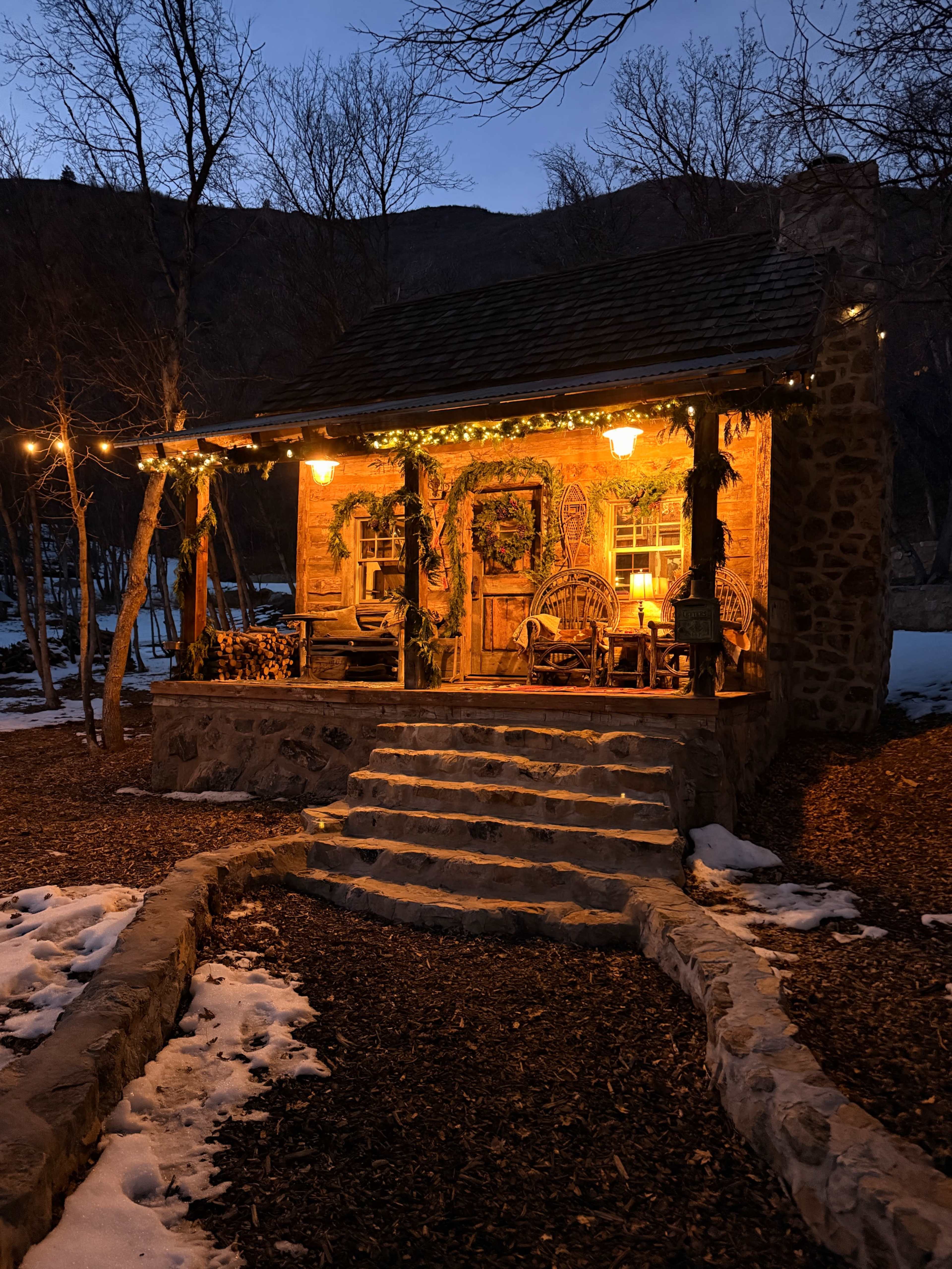 A stone cabin with warm lights and wooden rocking chairs sits at dusk, surrounded by bare trees and remnants of snow.