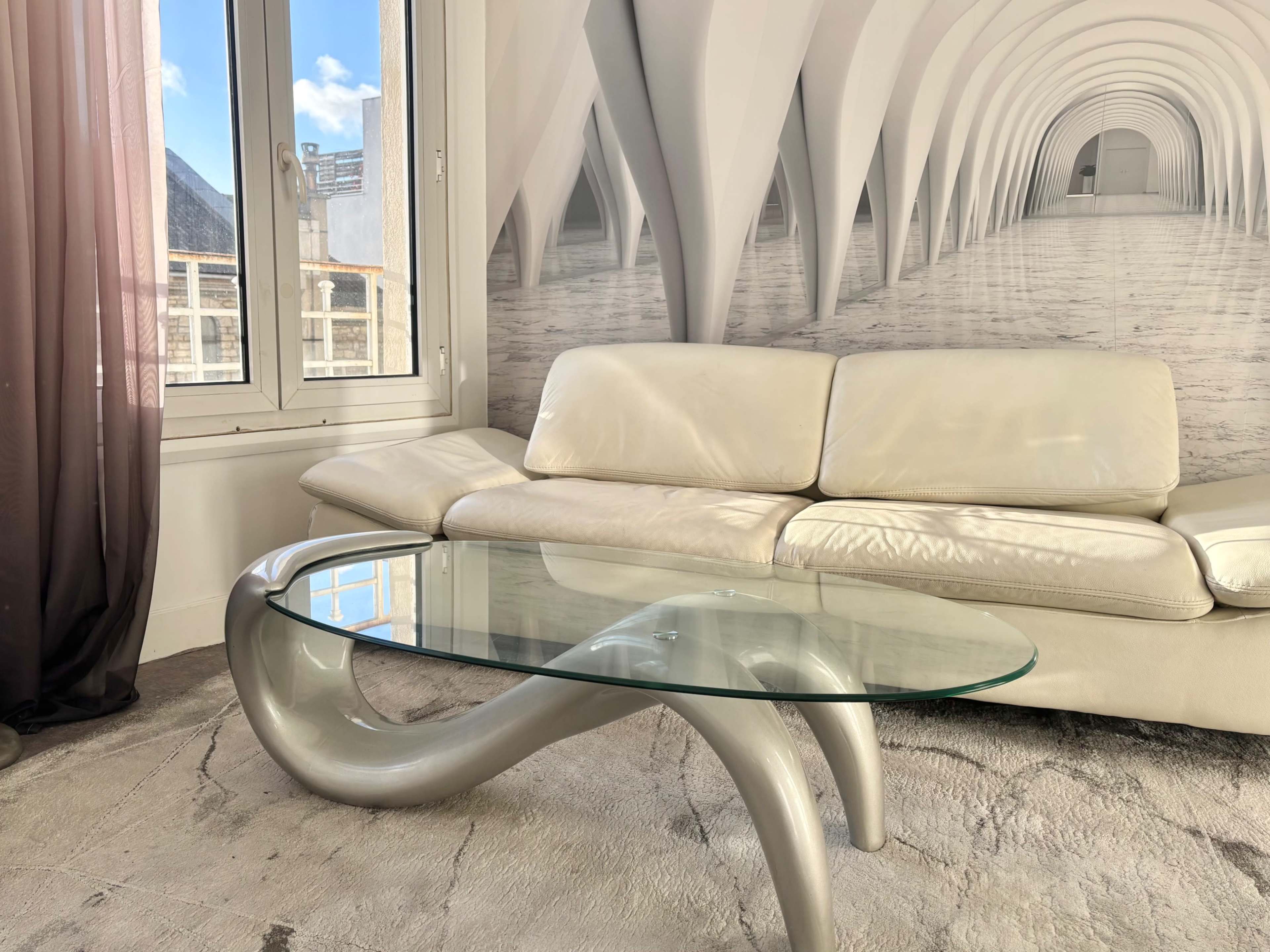 The image features a modern white sofa and a glass coffee table set against a backdrop of an arched architectural mural in a well-lit room.