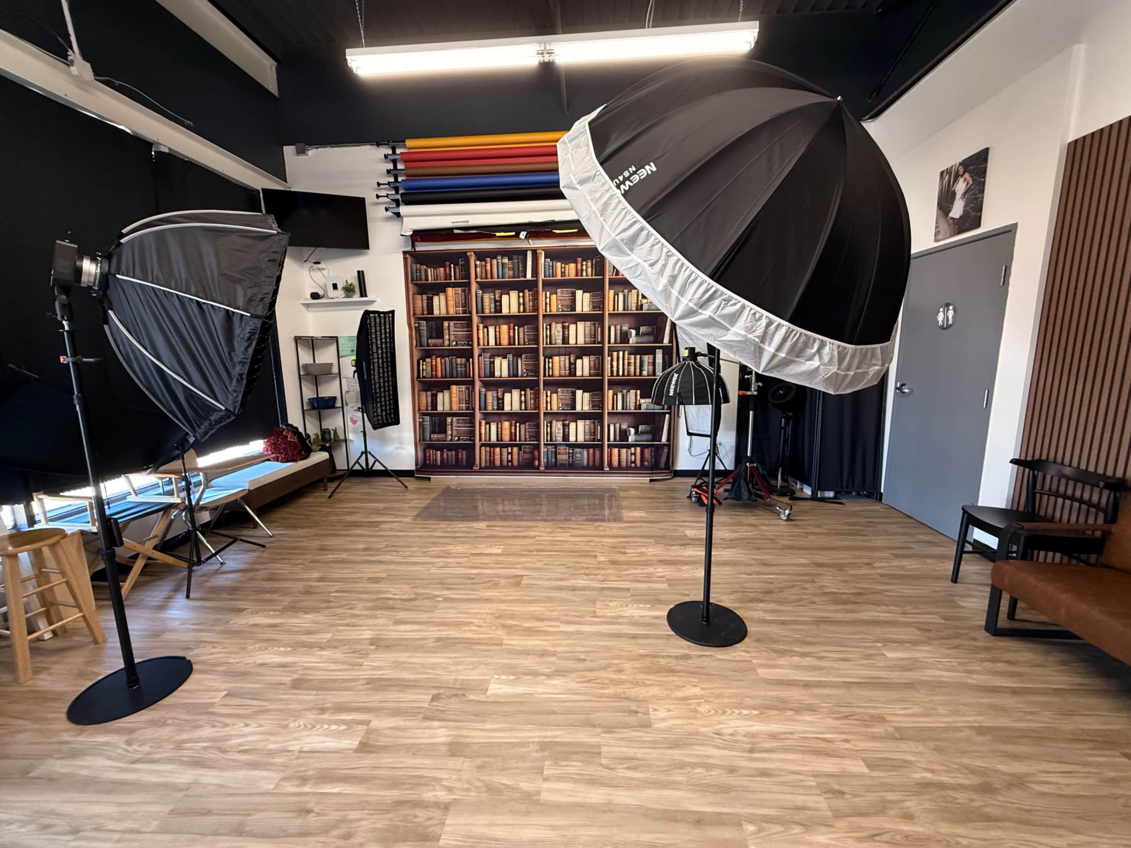 The image shows a photography studio featuring two large light umbrellas, a bookshelf backdrop, and various equipment arranged in the space.