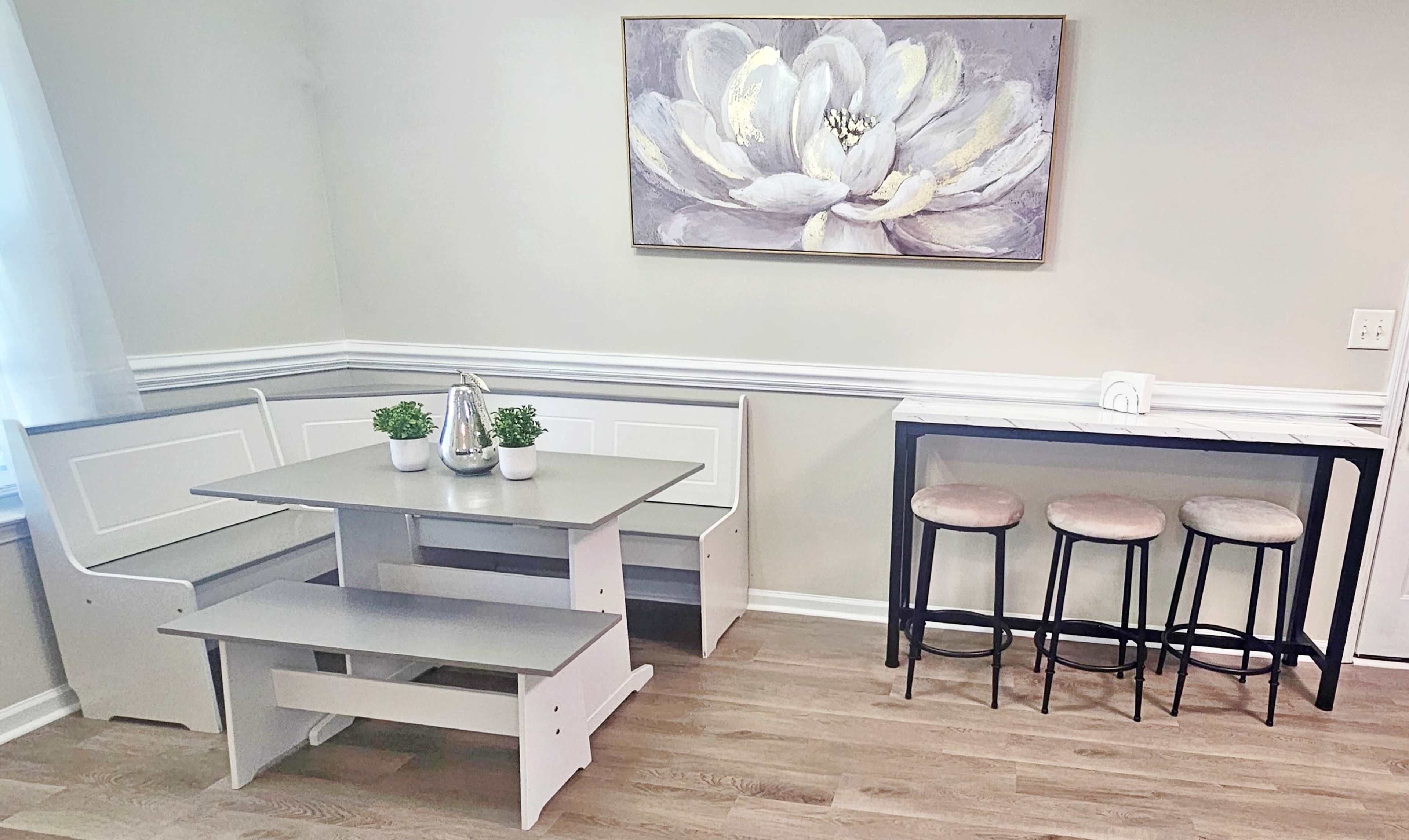 The image depicts a cozy dining area featuring a corner banquette with a table and two benches, alongside a narrow bar with three stools, all set against a light-colored wall adorned with a floral painting.