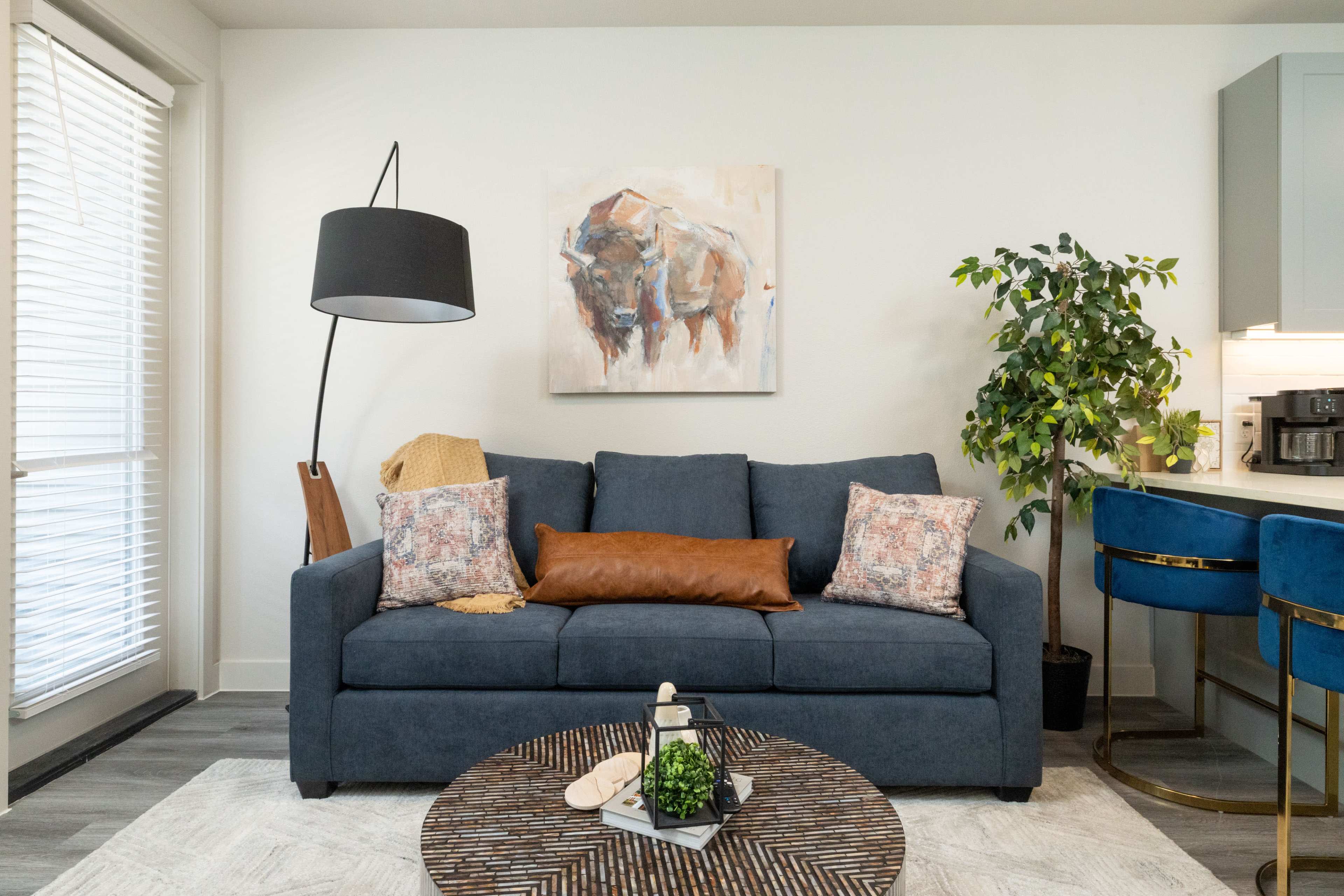 The image features a living room with a blue couch, decorative pillows, a round coffee table, and a tall plant beside a lamp.